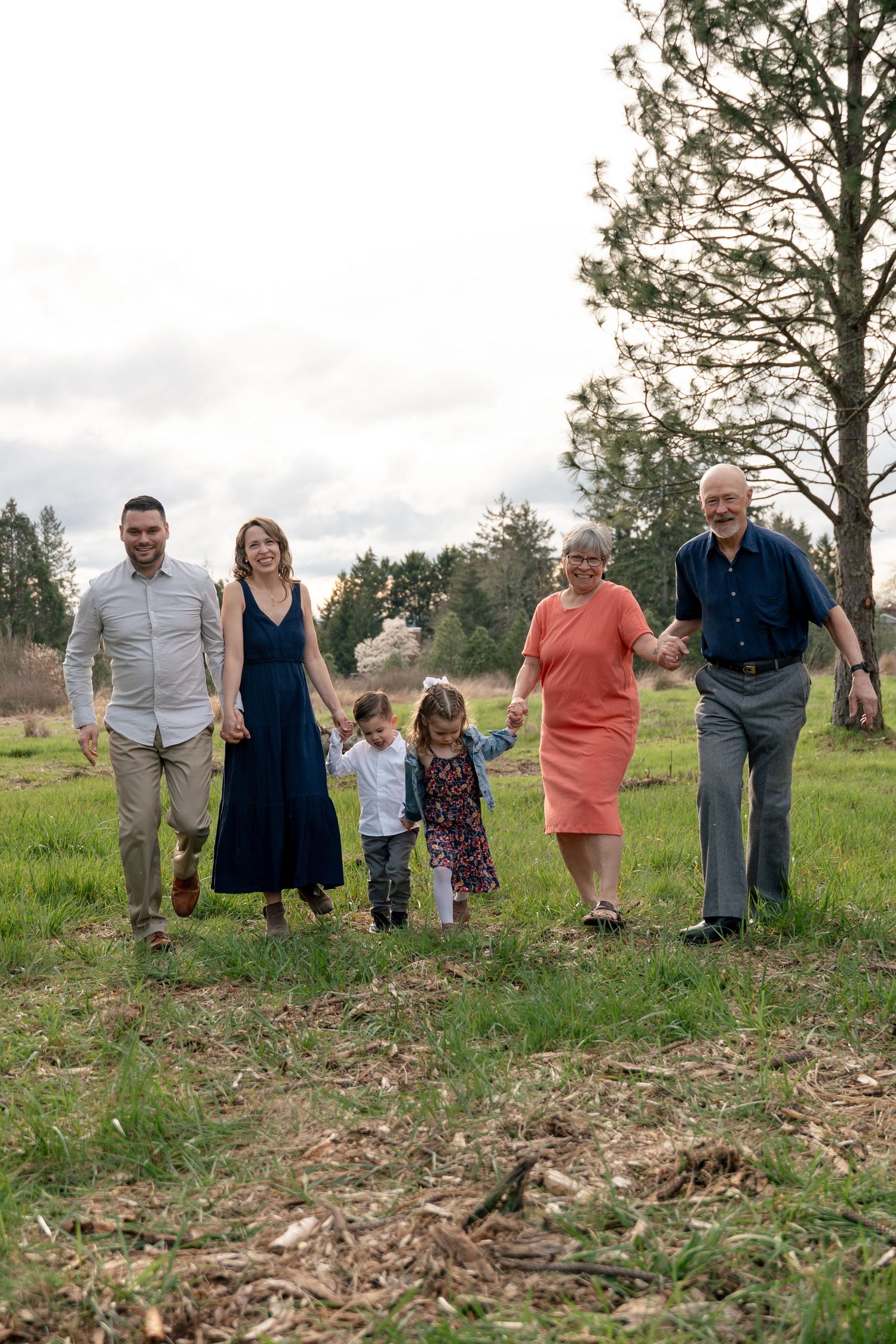 Multi-generational family of six walking hand-in-hand through green meadow on cloudy day.