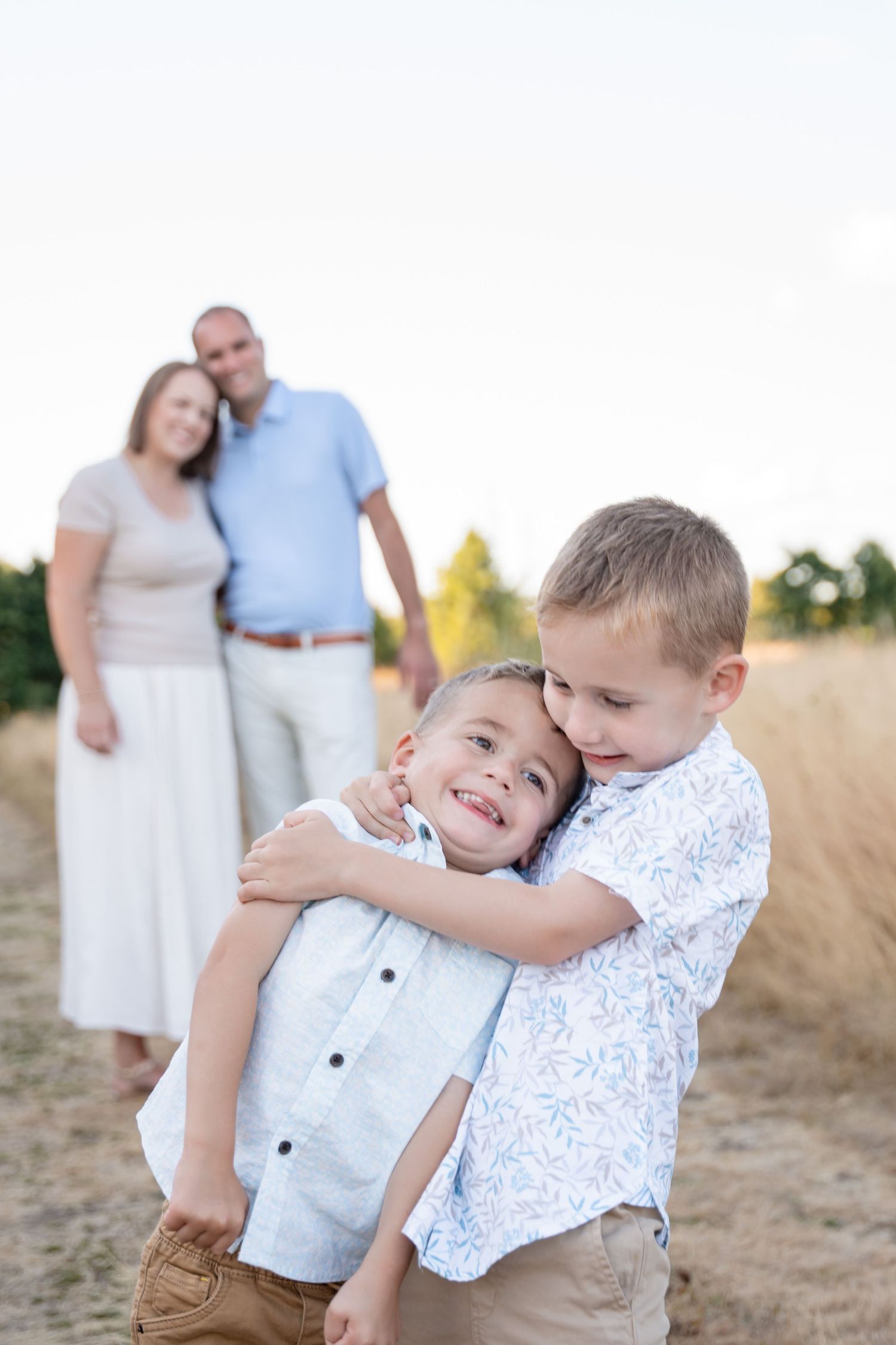An older brother hugs his younger sibling on a dirt path as parents smile behind them.