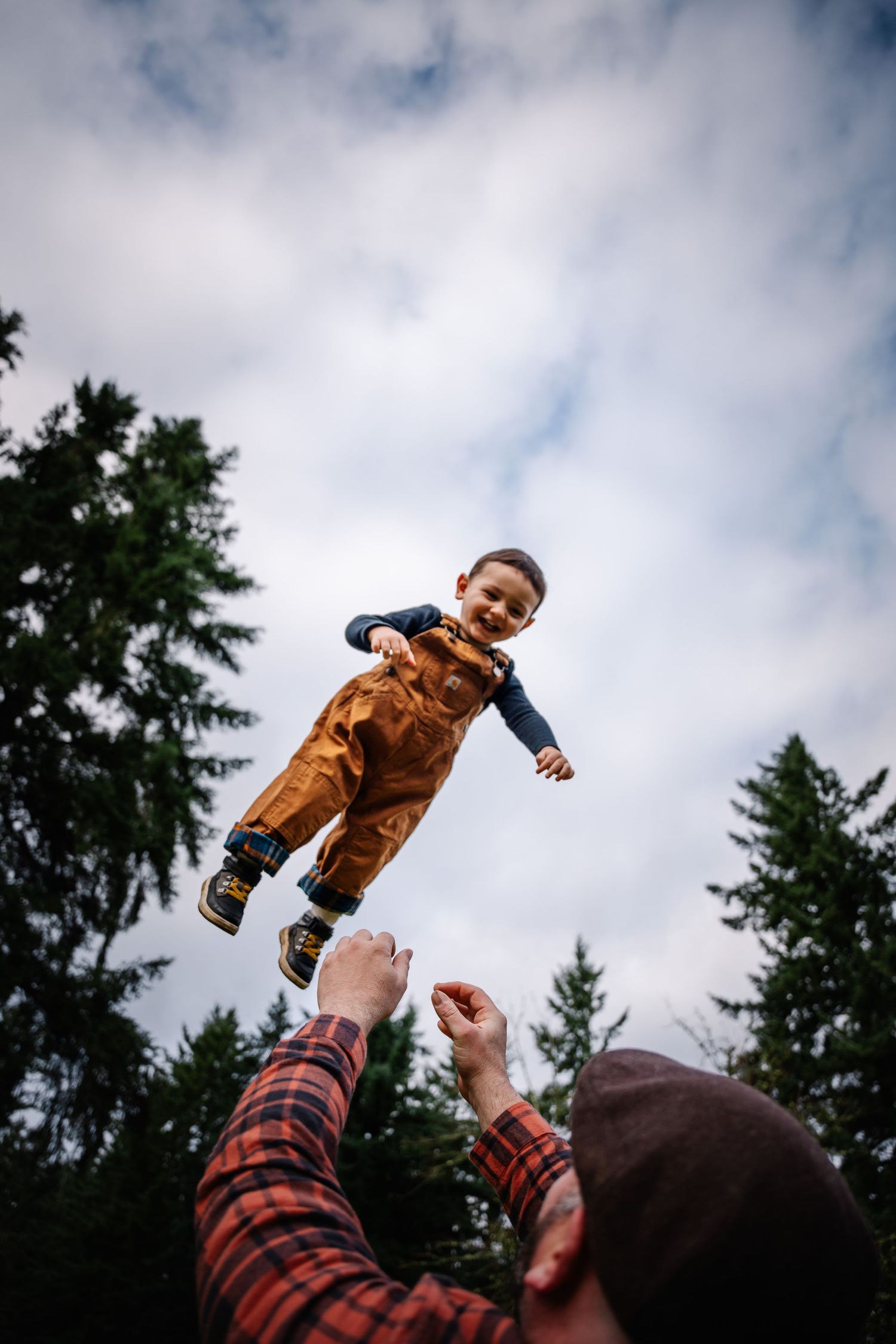 A joyful toddler in brown overalls is tossed into the air by a parent outdoors among tall evergreen trees.
