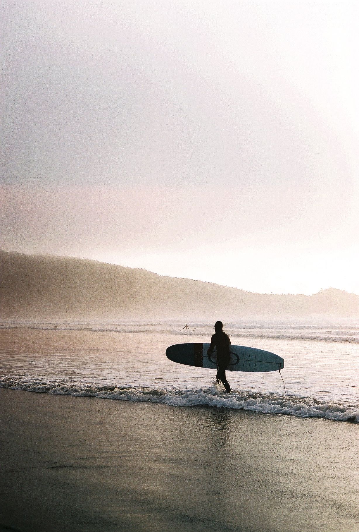 Silhouetted surfers walking along misty beach at sunset with surfboards in hand.