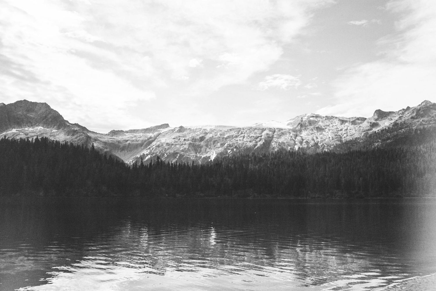 A serene black and white landscape of a mountain lake reflecting snow-capped peaks and forested shores.