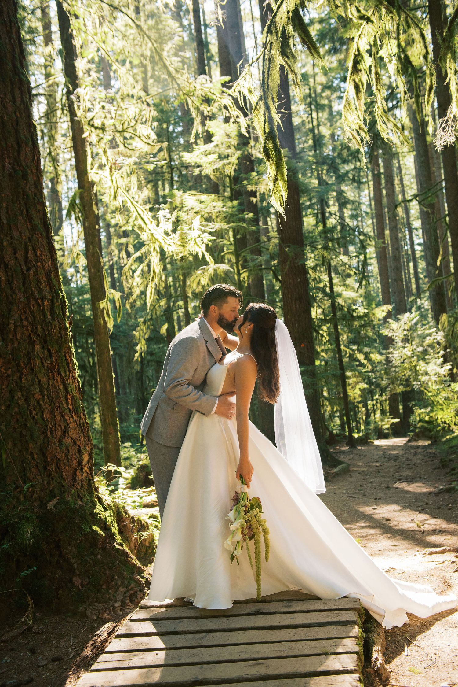 A romantic wedding couple shares a tender moment on a wooden boardwalk in a sunlit forest setting.