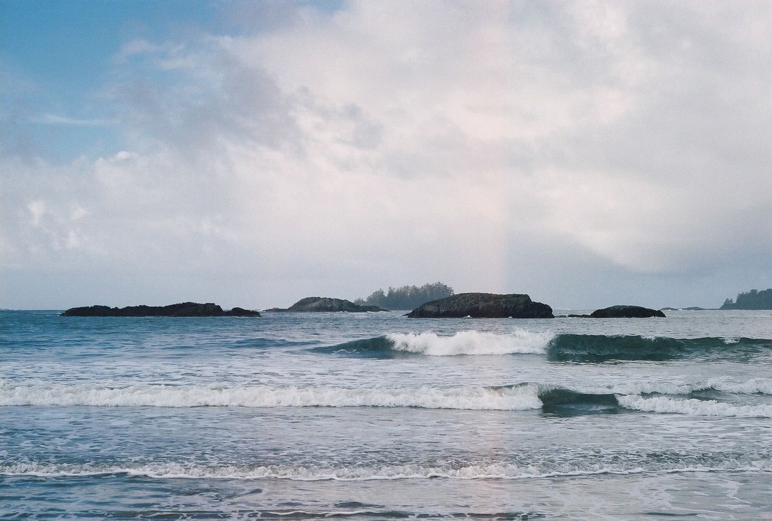Ocean waves roll towards shore with rocky islands visible on the horizon under a cloudy blue sky.