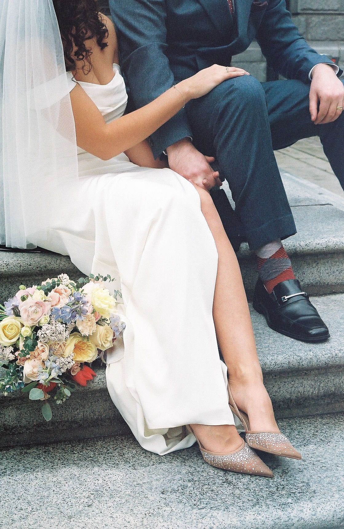 Close-up shot of bride and groom sitting on stone steps with floral bouquet.