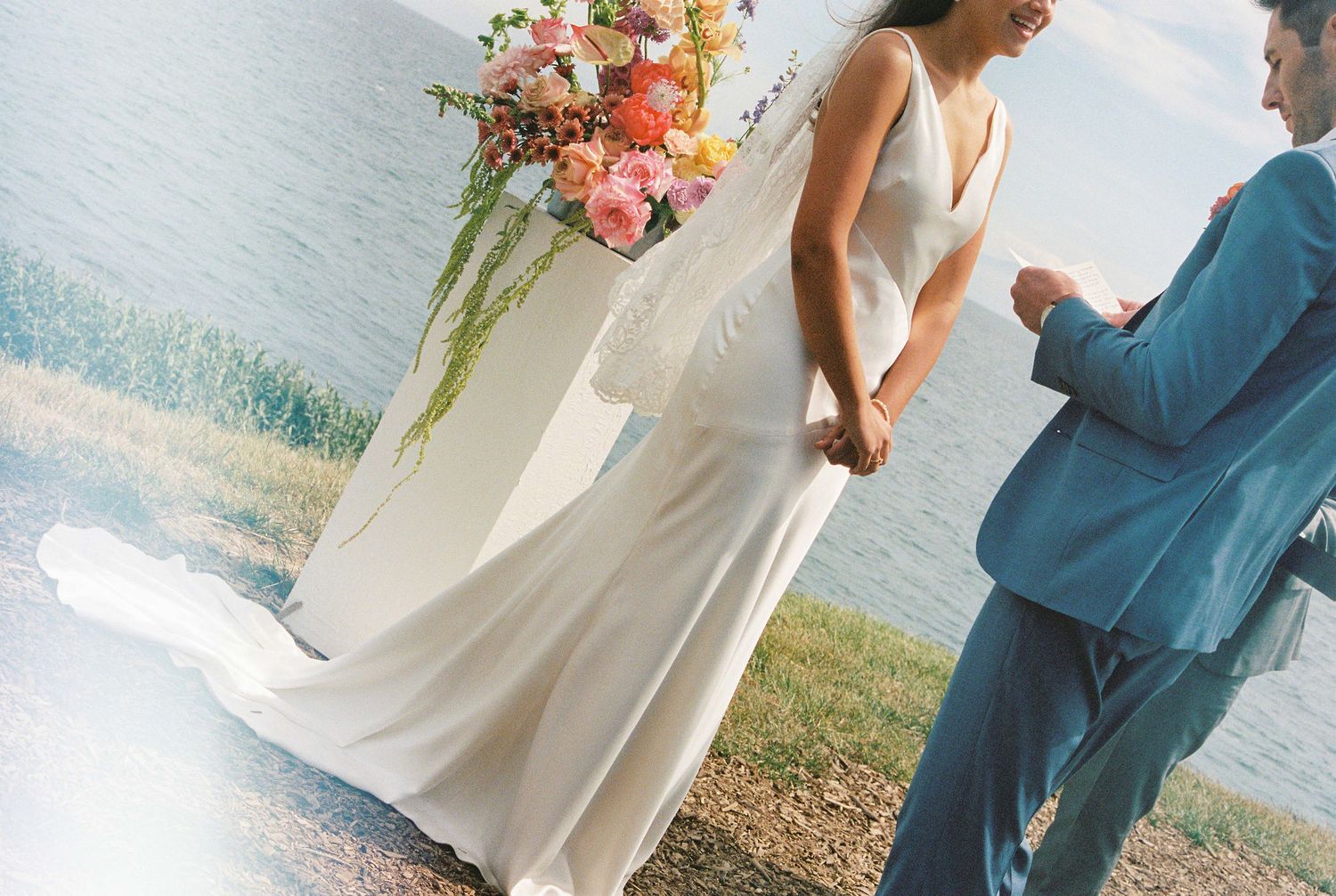 Bride in white gown and groom in teal suit exchange vows outdoors by water with colorful floral arrangement.