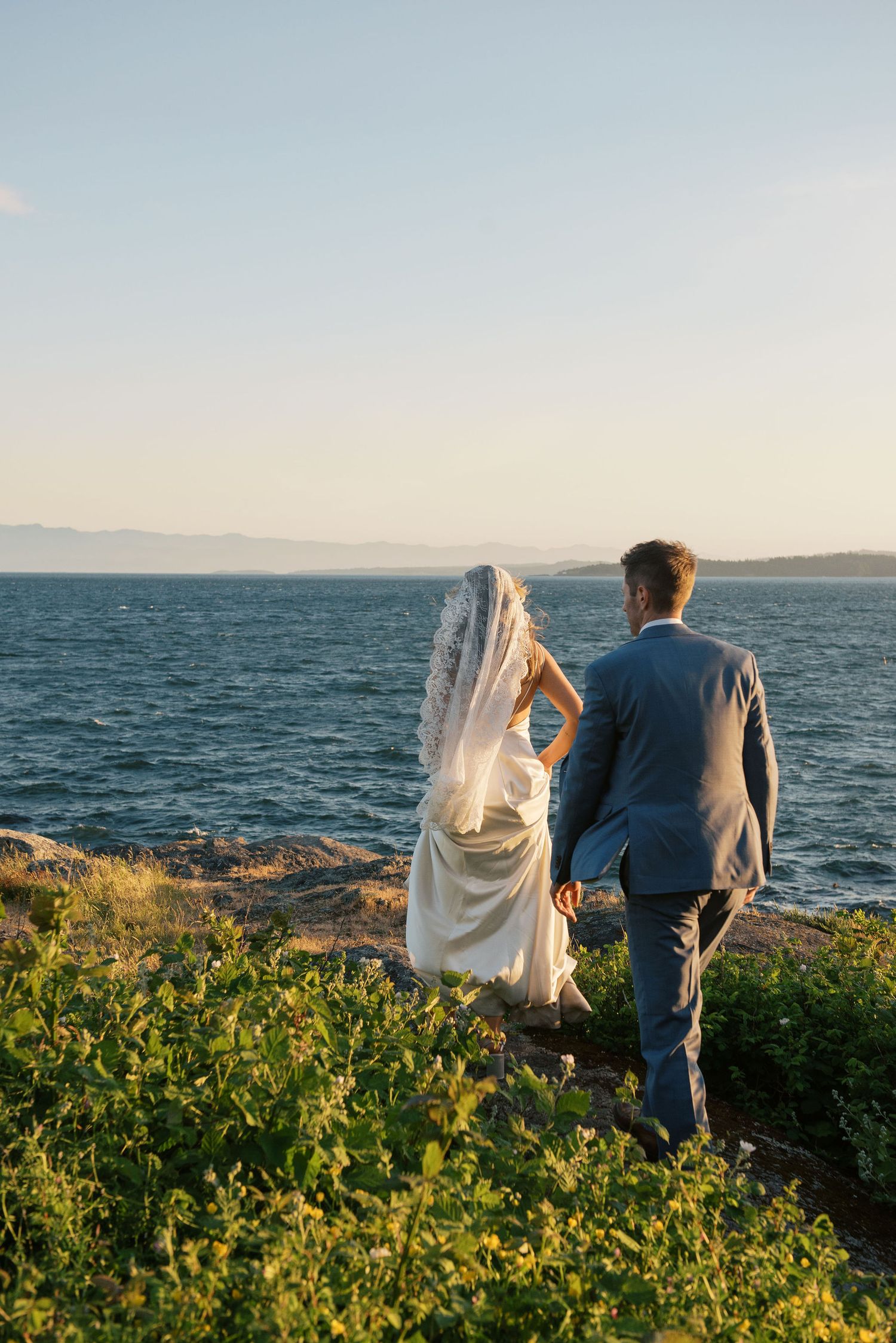 Bride and groom holding hands facing the ocean at sunset, bride wearing white gown and veil.