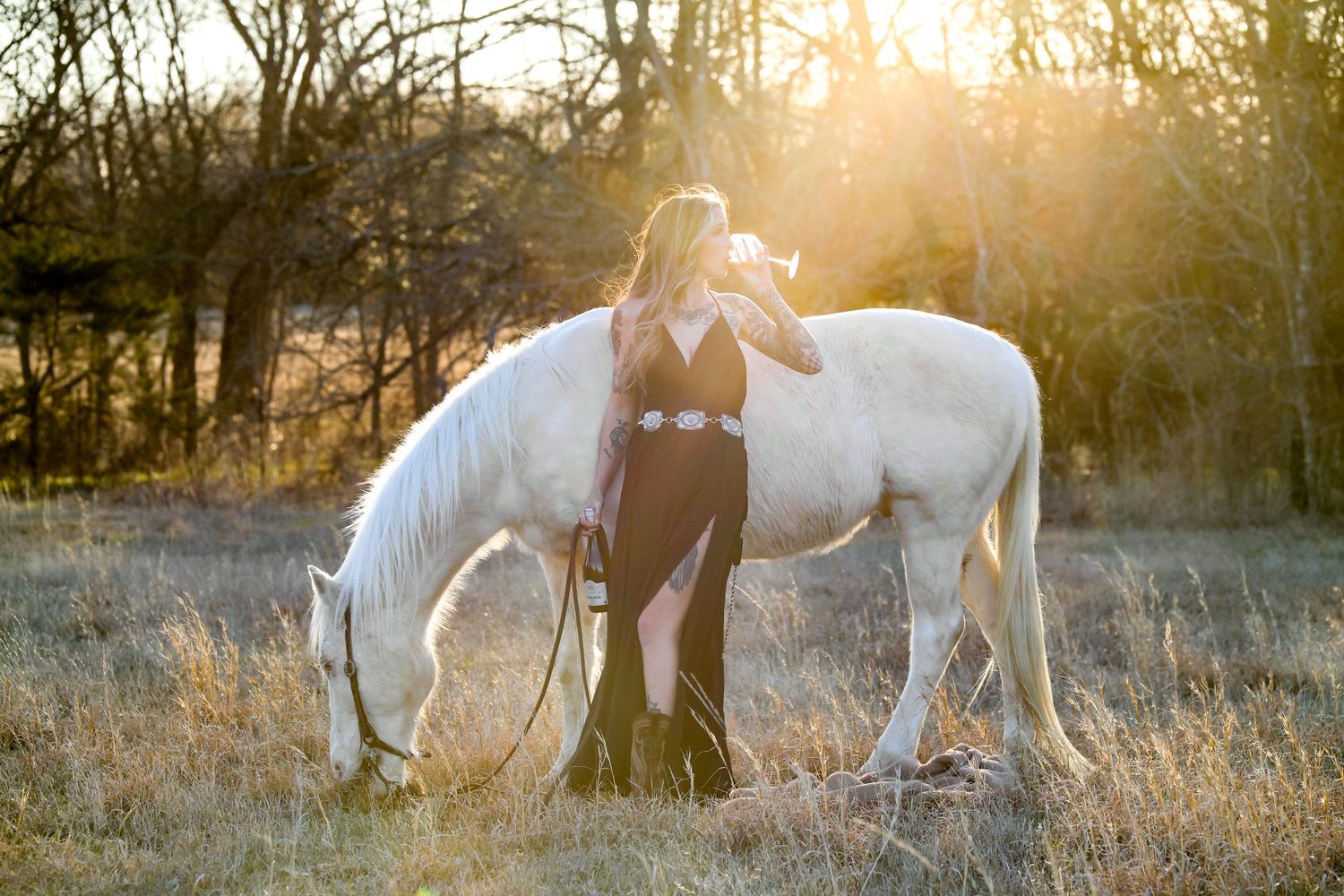 girl wearing a black dress sipping a glass of wine standing next to a white horse in a field at sunset. luxury equestrian pictures