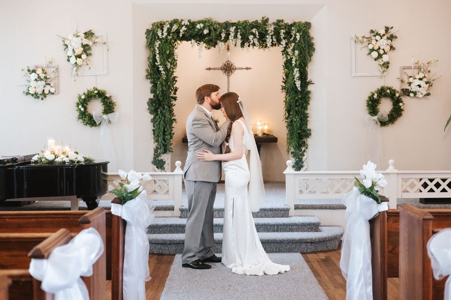 Indoor wedding ceremony setup with white floral arch and greenery against a light colored wall.