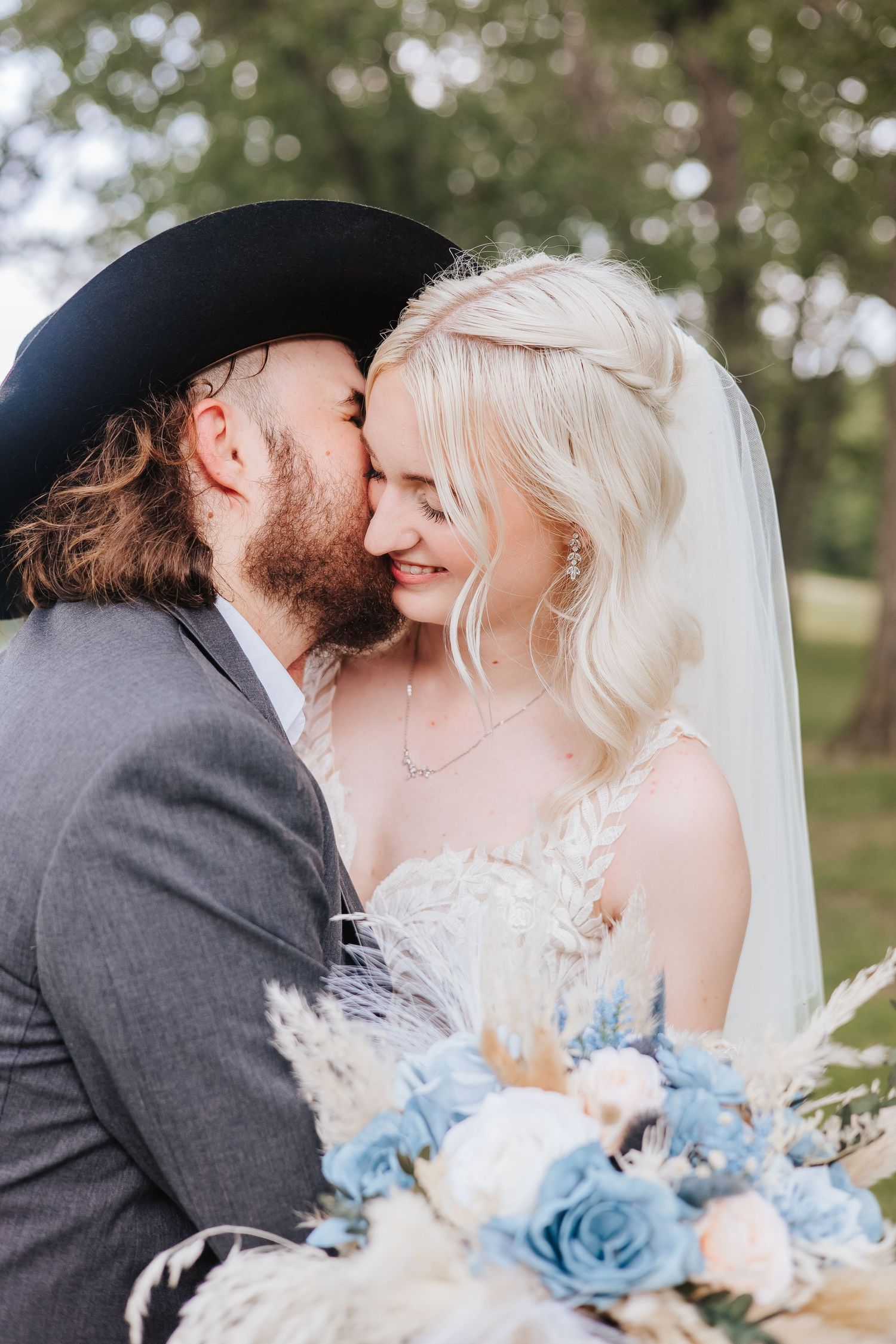 Romantic wedding portrait with blue bouquet and cowboy hat against natural outdoor backdrop.