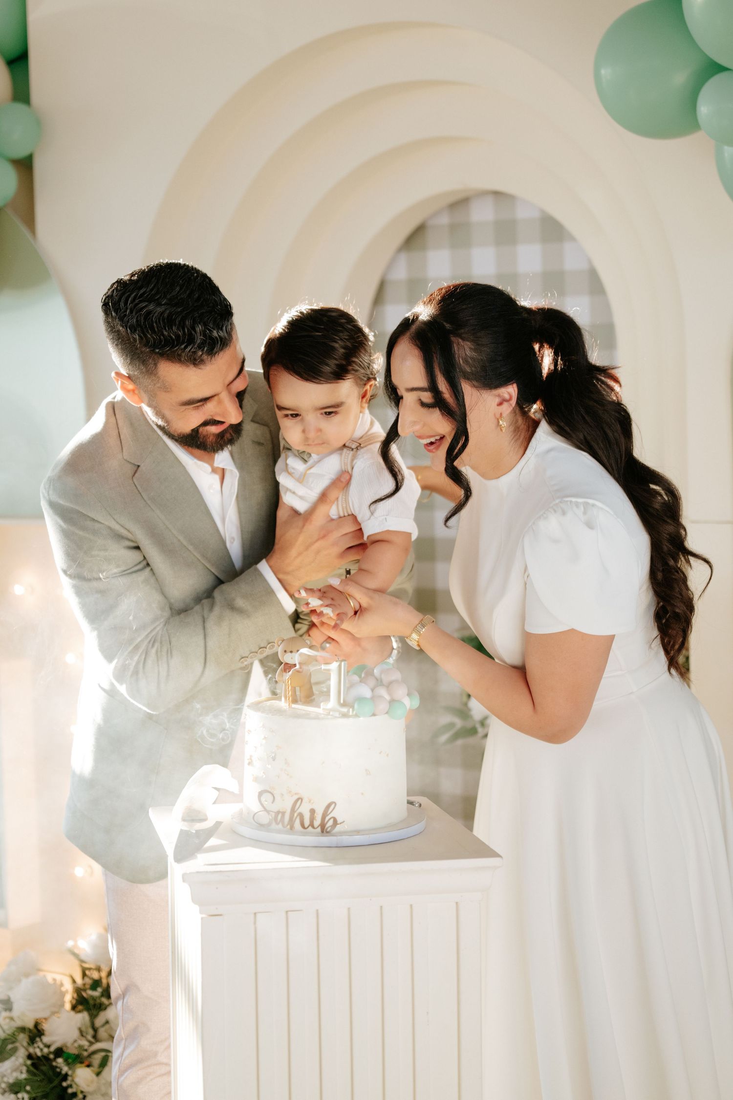 Family celebrating a baby’s first birthday, cutting a white cake together at a decorated party setup with green balloons.