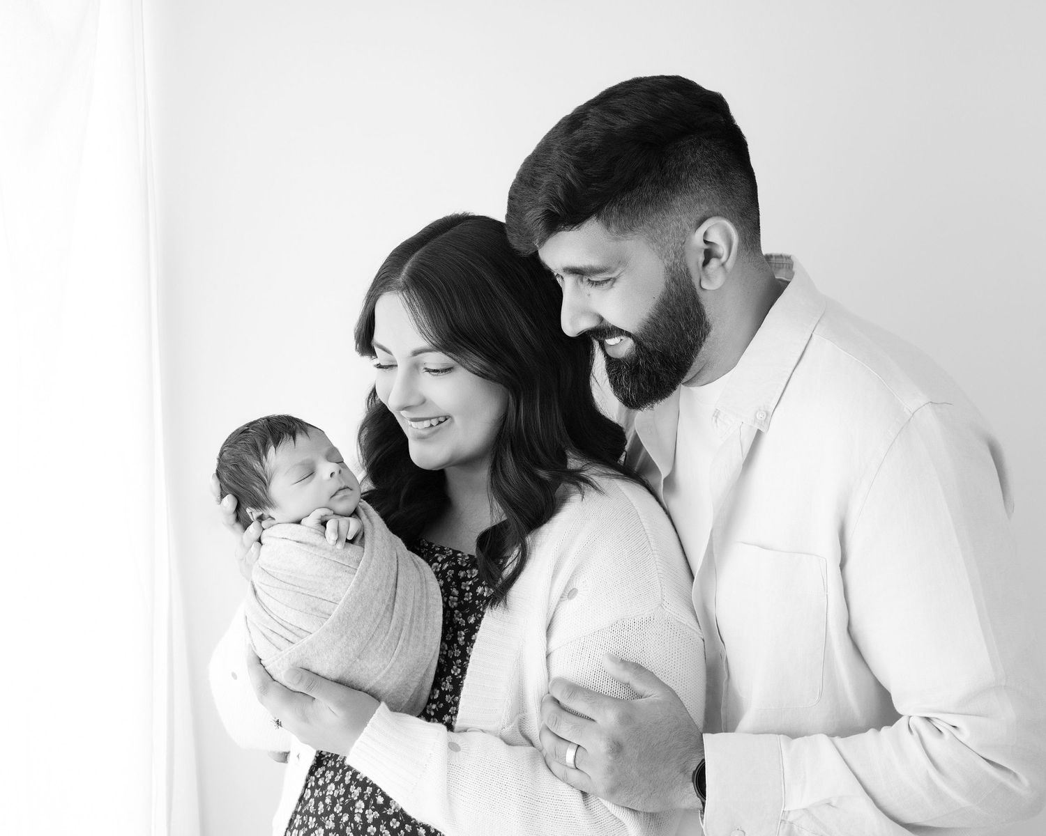 Black and white newborn family portrait of smiling parents holding their swaddled baby against a bright white background.