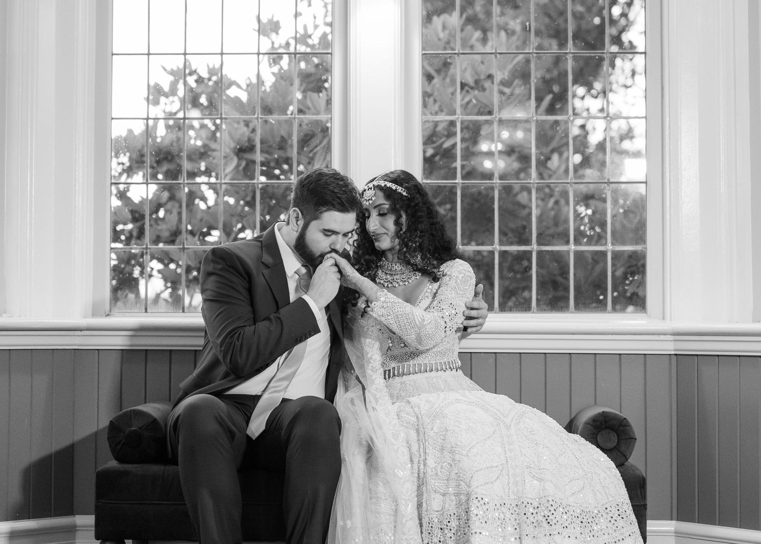 Black and white wedding photo of groom kissing bride's hand while sitting together near large window.