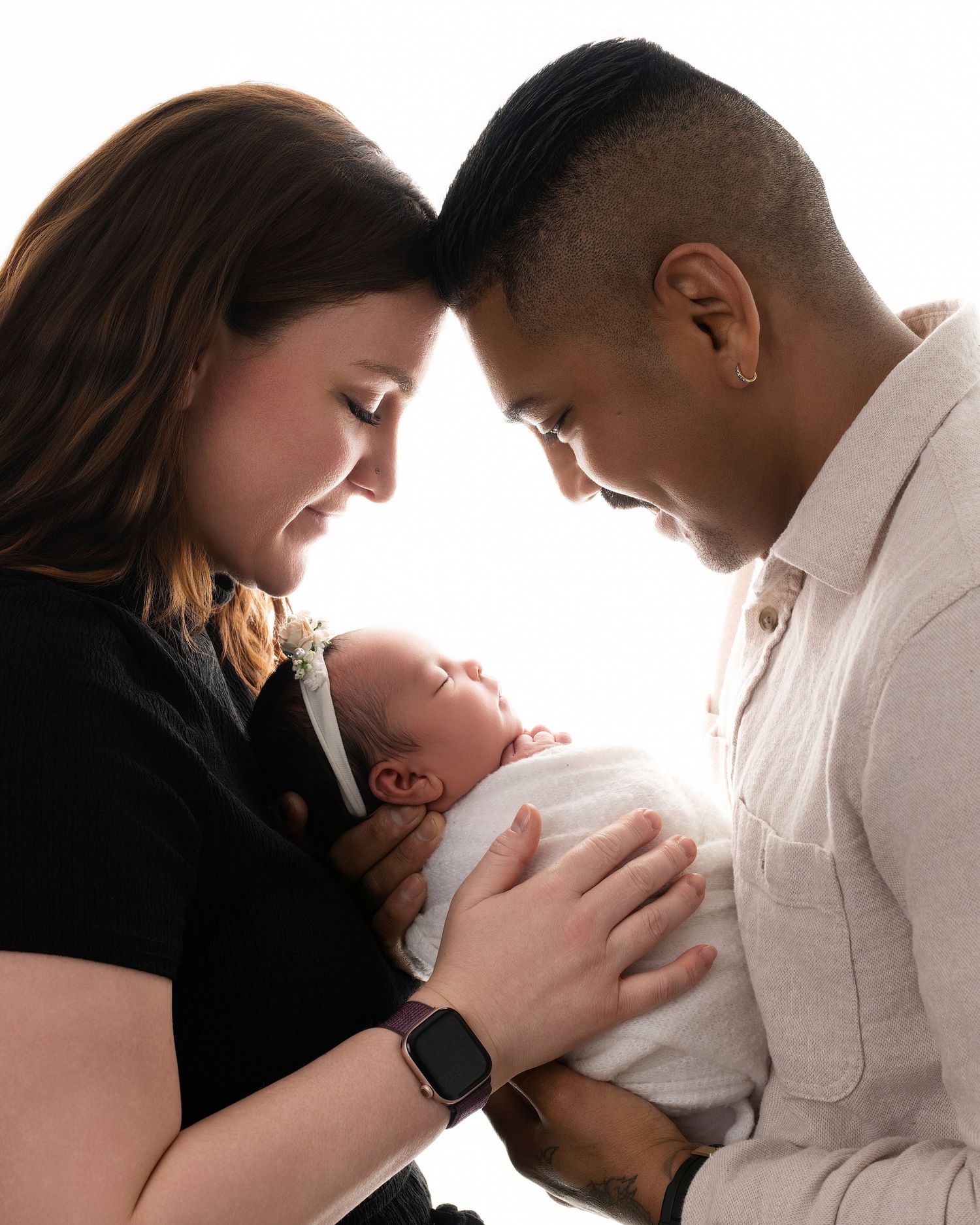 New parents tenderly cradle their newborn baby together, foreheads touching in an intimate studio portrait.