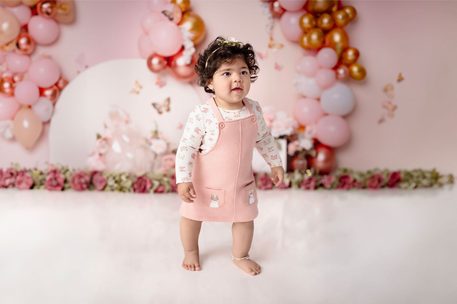 Baby girl in pink dress stands before balloon arch backdrop during first birthday cake smash photo session.