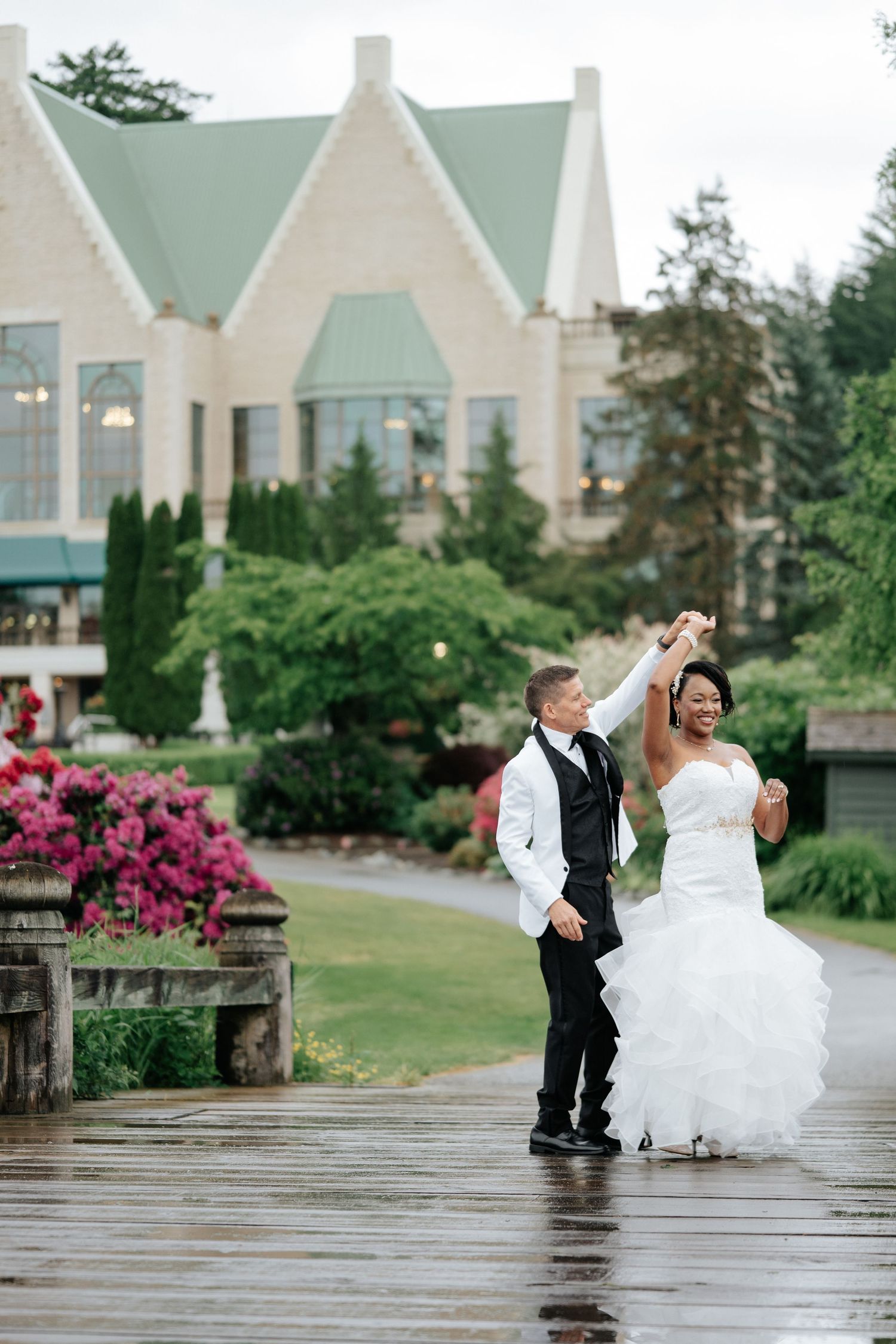 Bride and groom dancing on a rainy outdoor pathway with a grand wedding venue and lush greenery in the background.