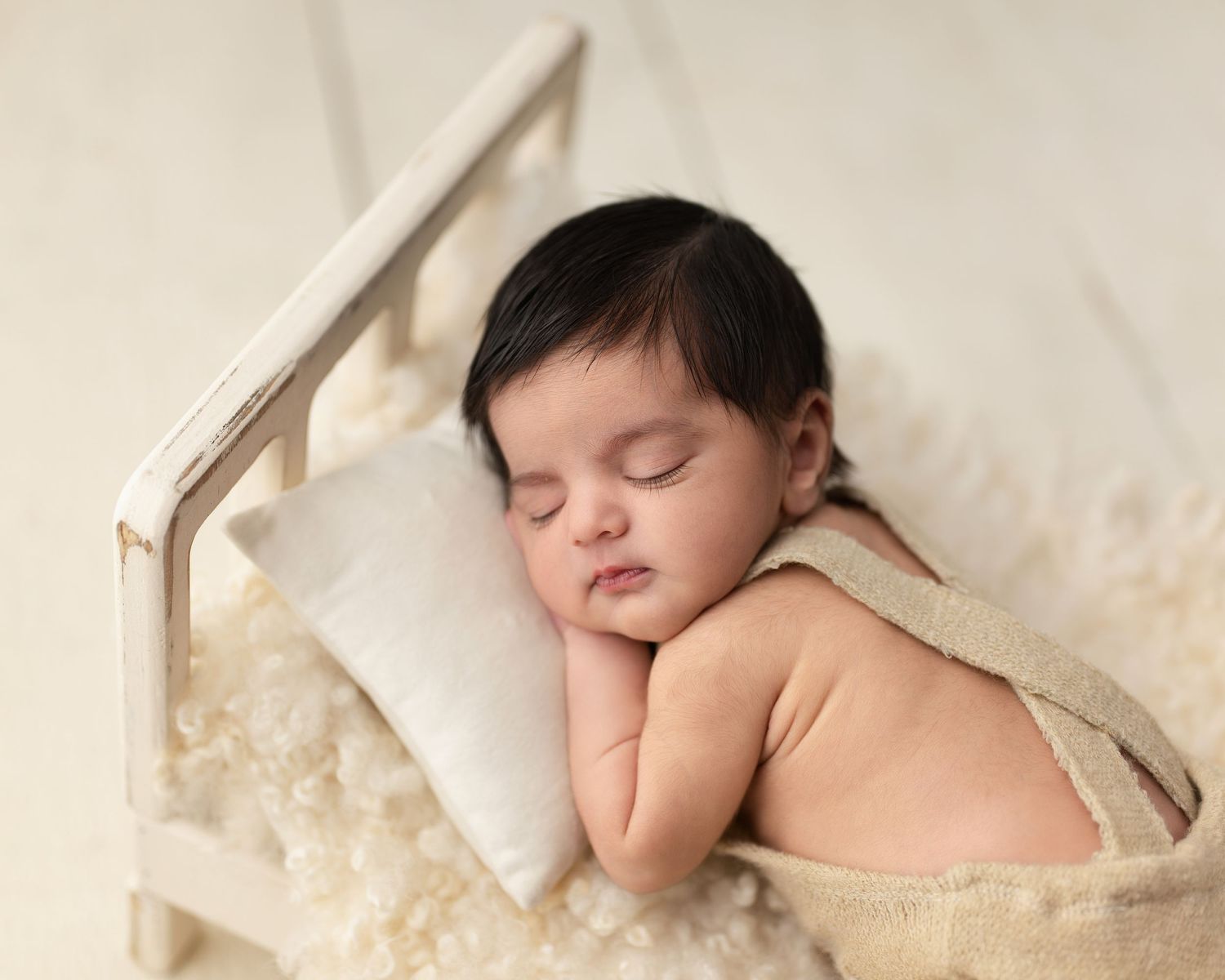 Peaceful newborn baby with dark hair sleeping on a tiny wooden bed with a white pillow and fluffy rug.
