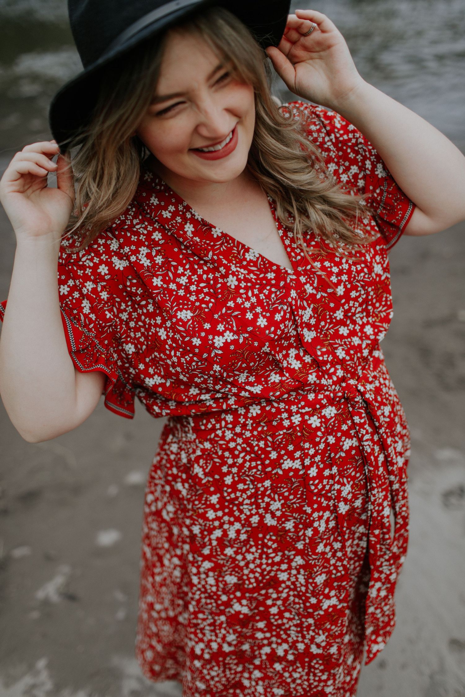 Person wearing red floral dress and black hat smiling during outdoor photoshoot.