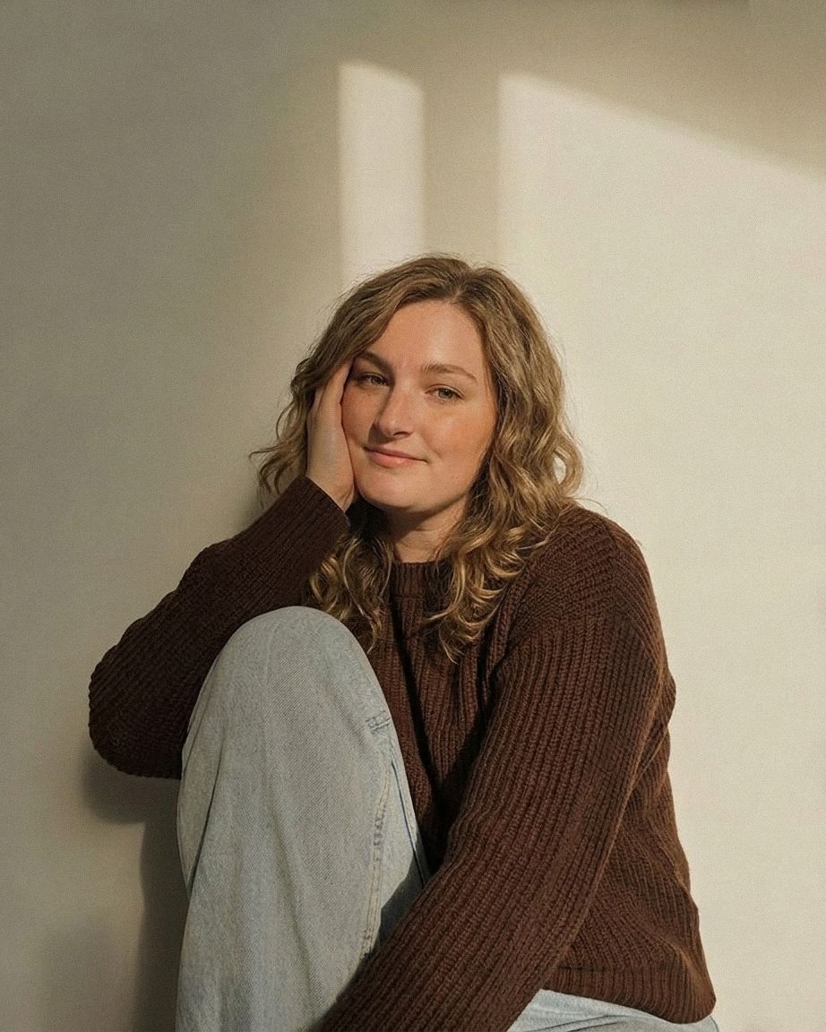 Young woman with curly blonde hair in brown knit sweater and jeans sitting relaxed against a sunlit white wall.