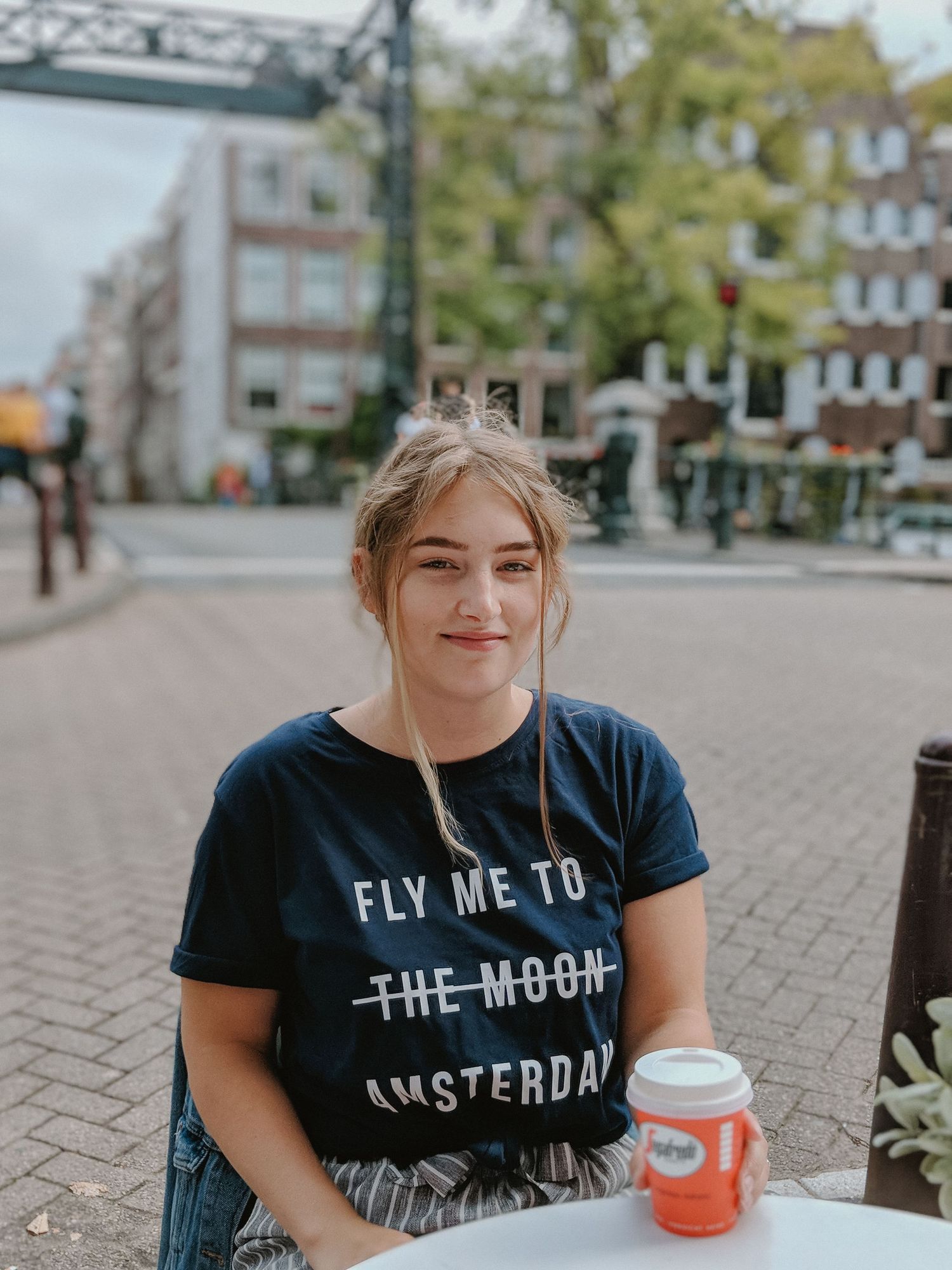 Person in navy t-shirt with 'Fly Me to Amsterdam' text sitting outdoors with beverage.