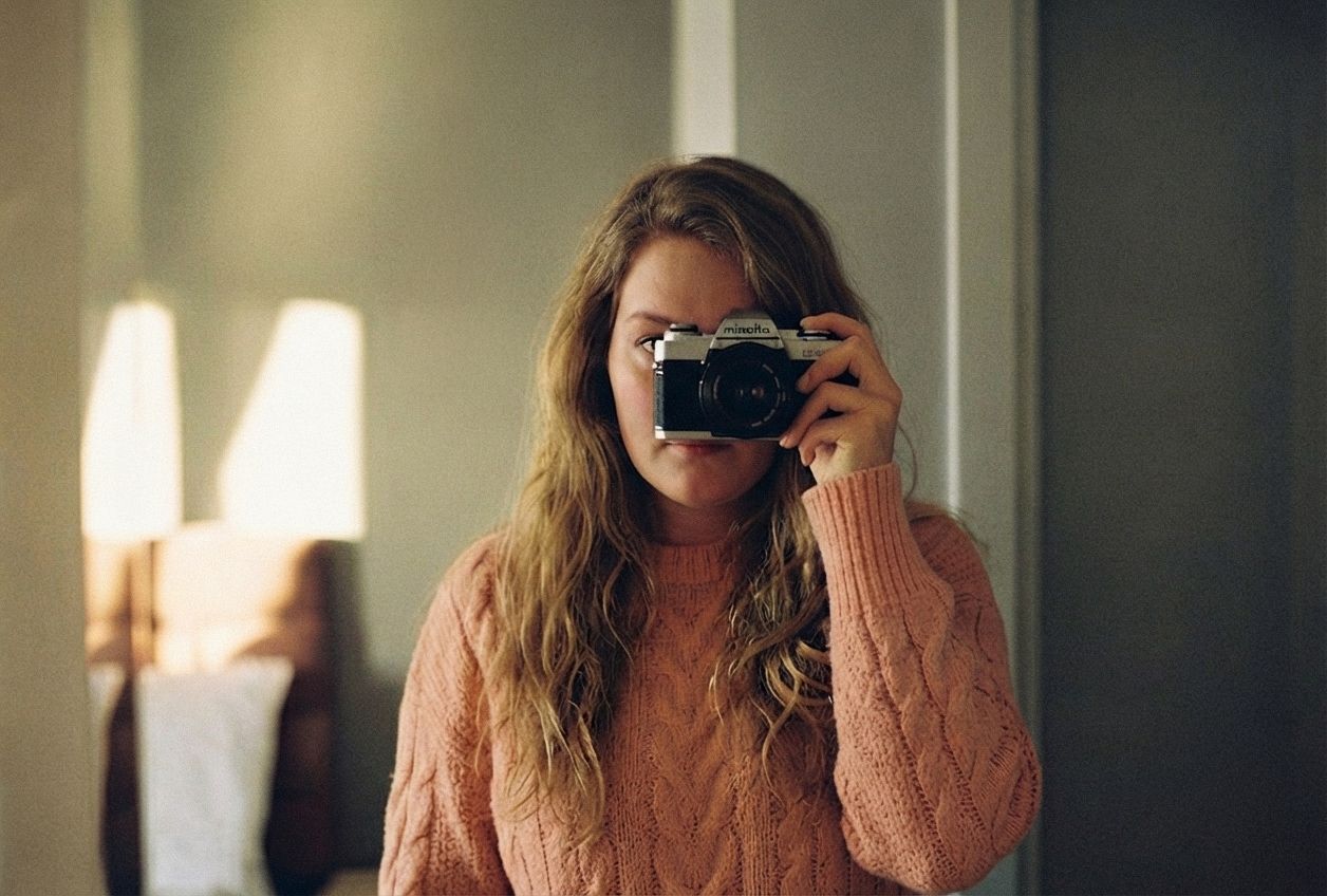 Young woman in pink cable-knit sweater taking a self-portrait with a vintage film camera in a mirror.