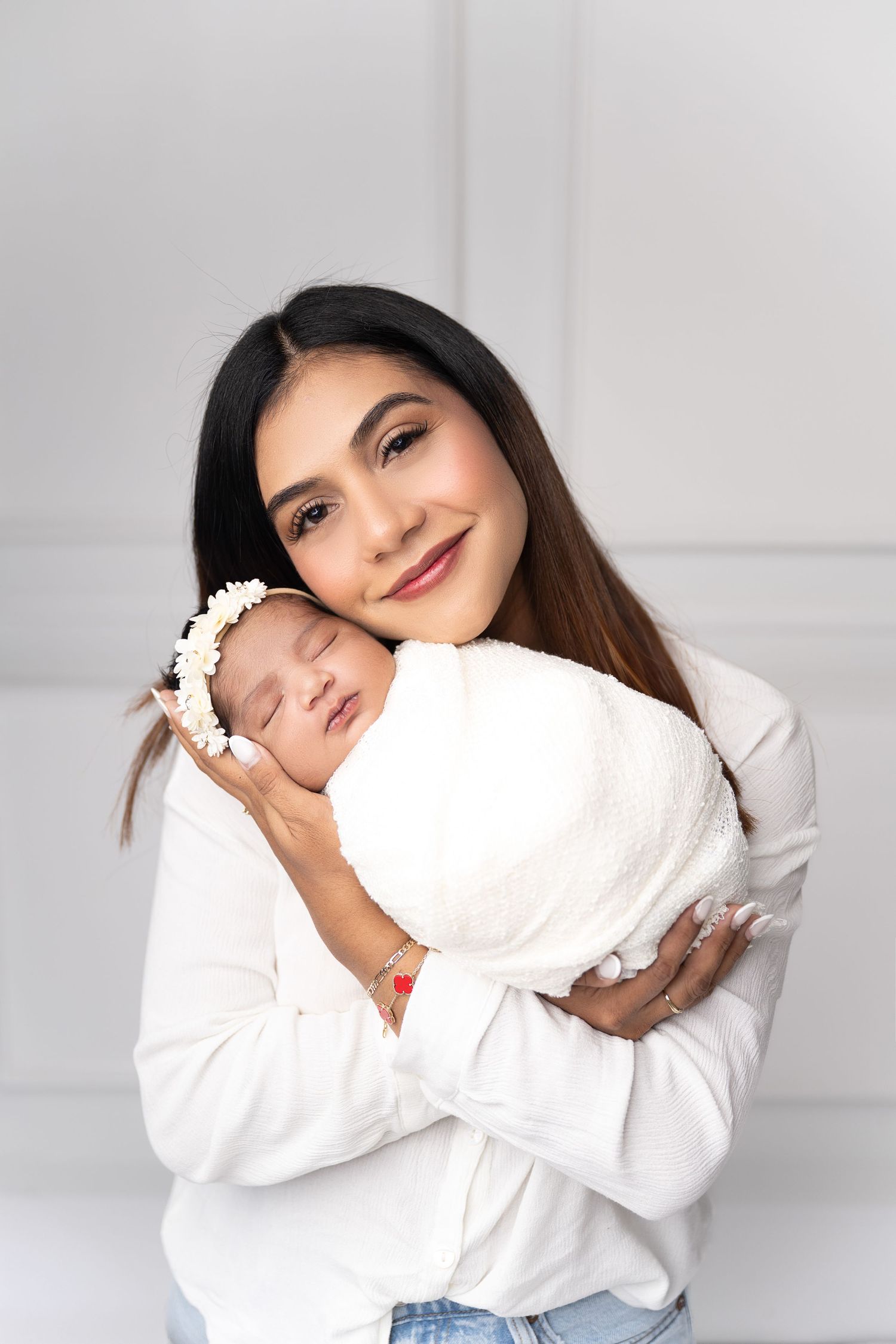 A smiling young mother in a white shirt tenderly holds her sleeping newborn baby wrapped in a white swaddle.