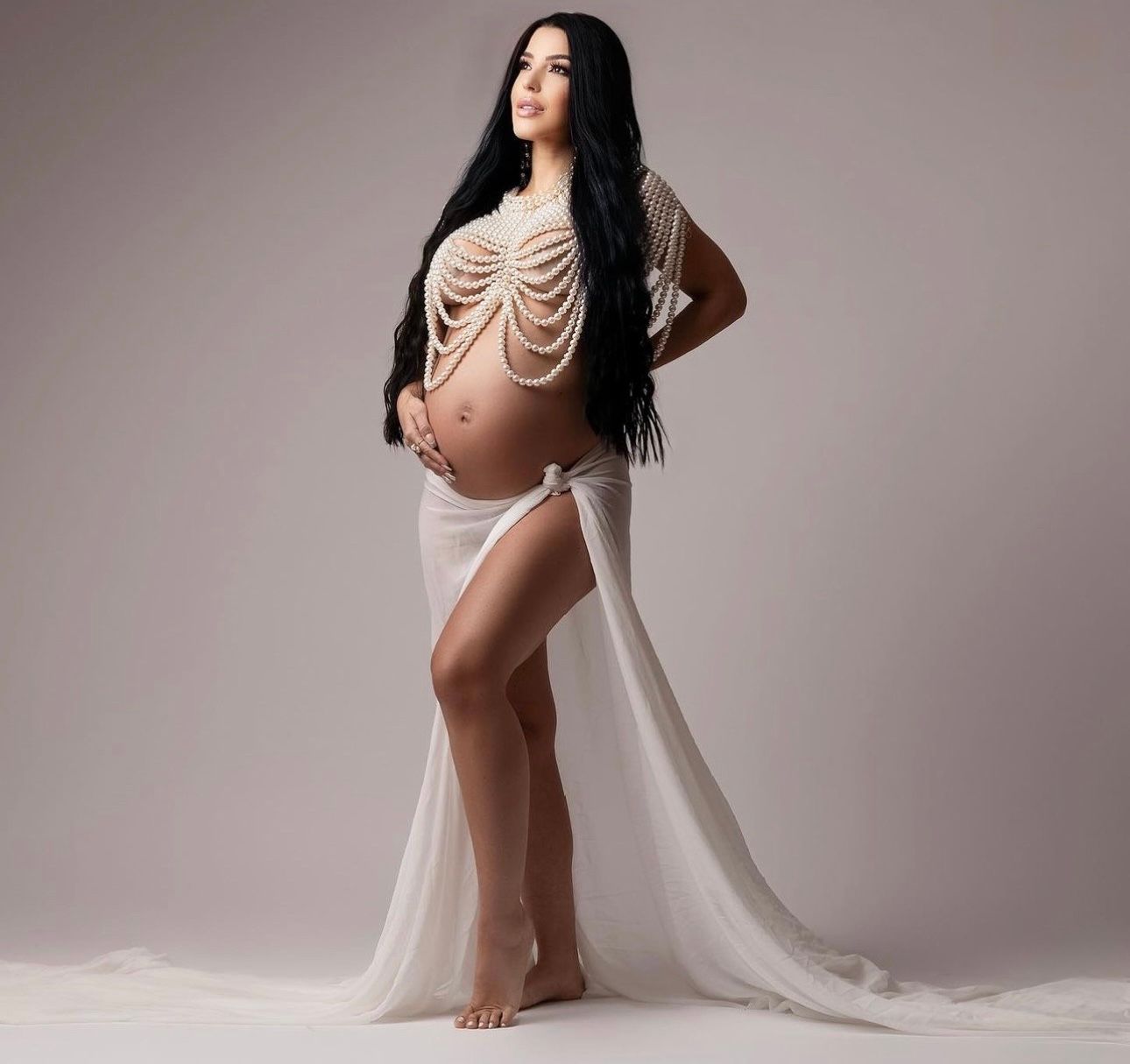 Pregnant woman in sheer white maternity gown with pearl body chain posing for professional maternity photo shoot.