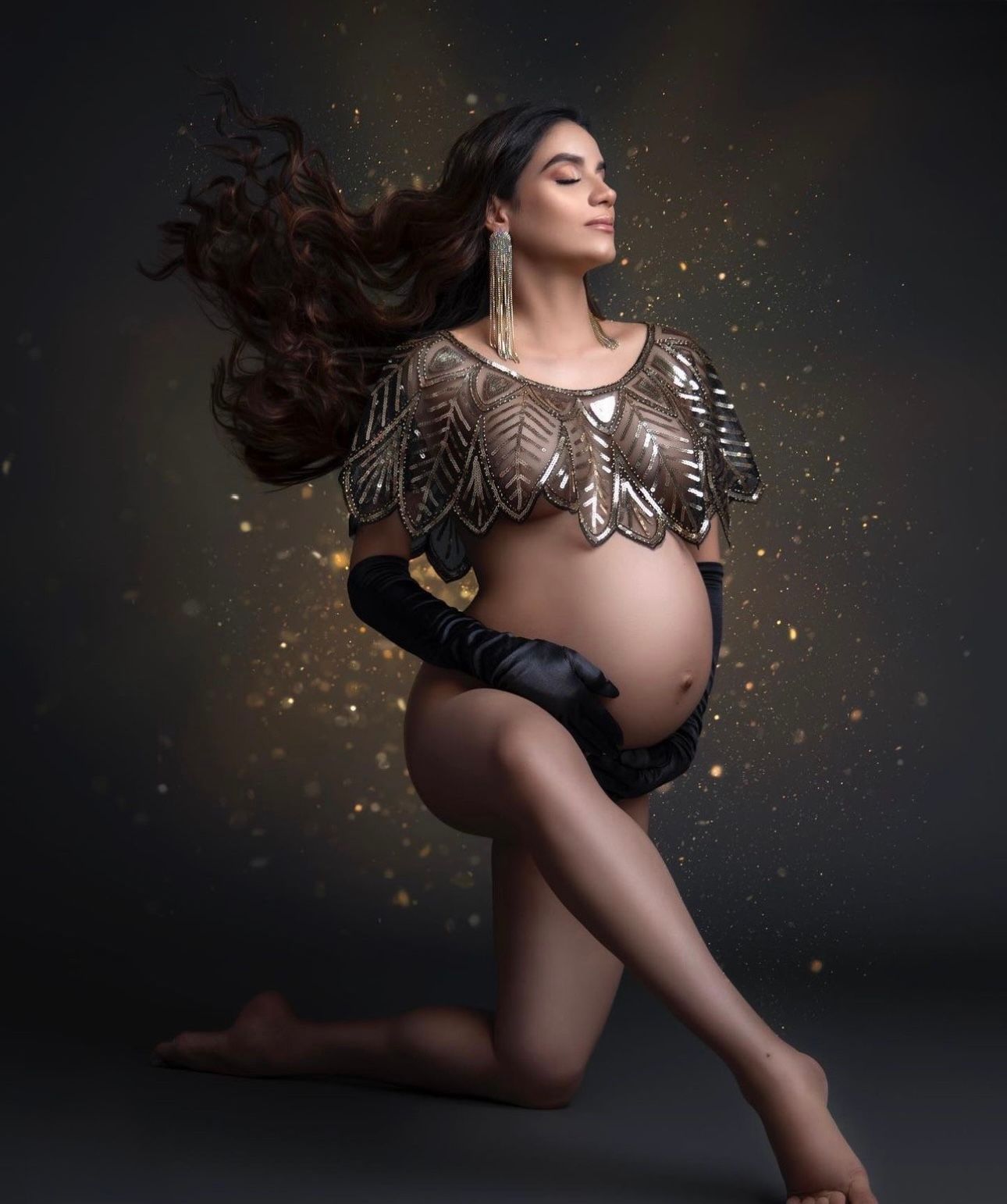 Pregnant woman in golden leaf armor top poses gracefully on knees with flowing hair and black gloves on dark background.