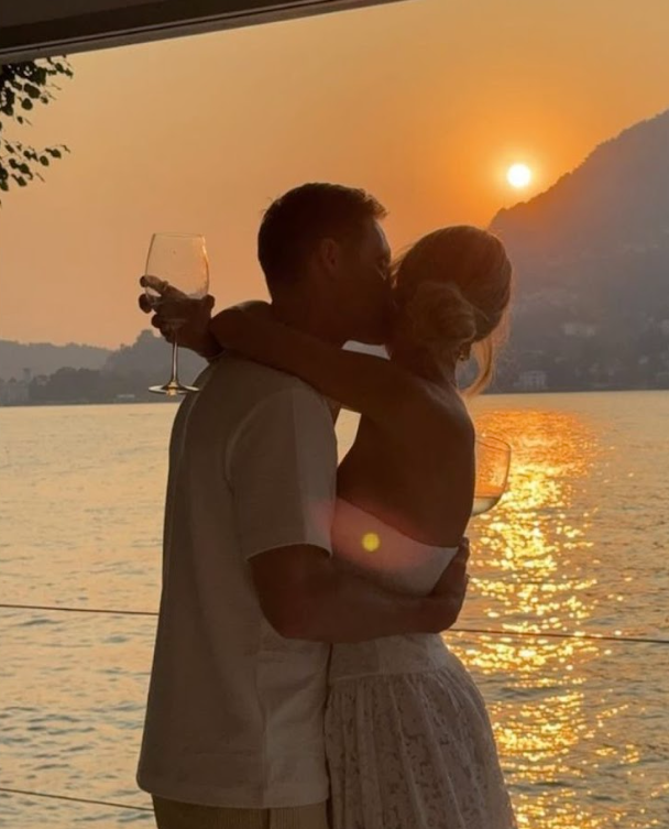 Silhouetted couple embraces while watching a golden sunset over calm waters with mountains in the distance.