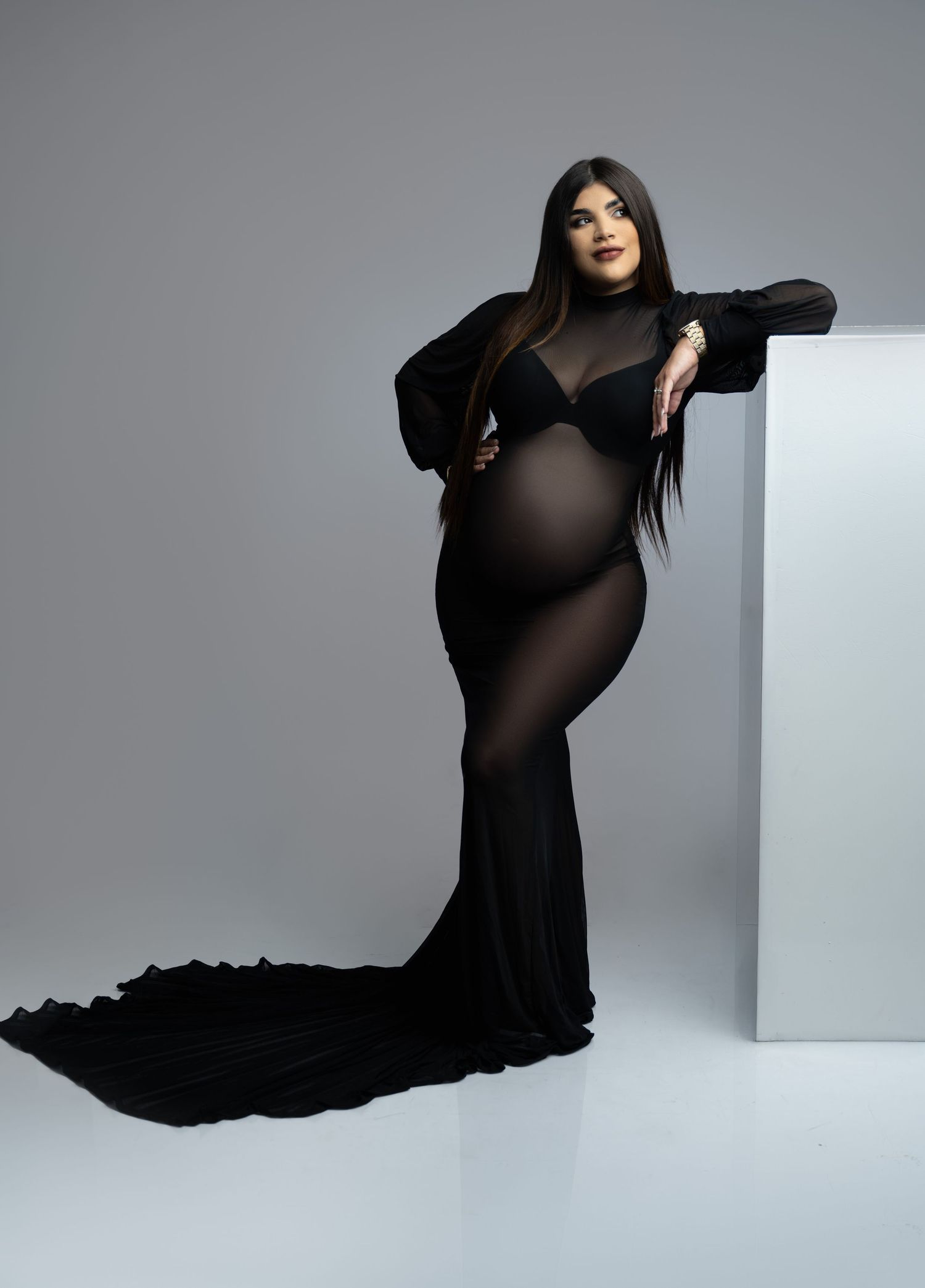 Elegant maternity photoshoot featuring a flowing black sheer gown with train against a minimalist gray backdrop.