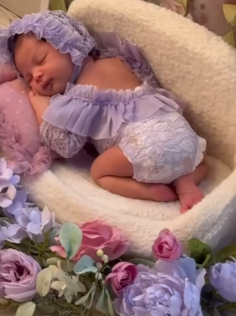 Baby in lavender dress sleeping peacefully among purple flowers on cream cushion.
