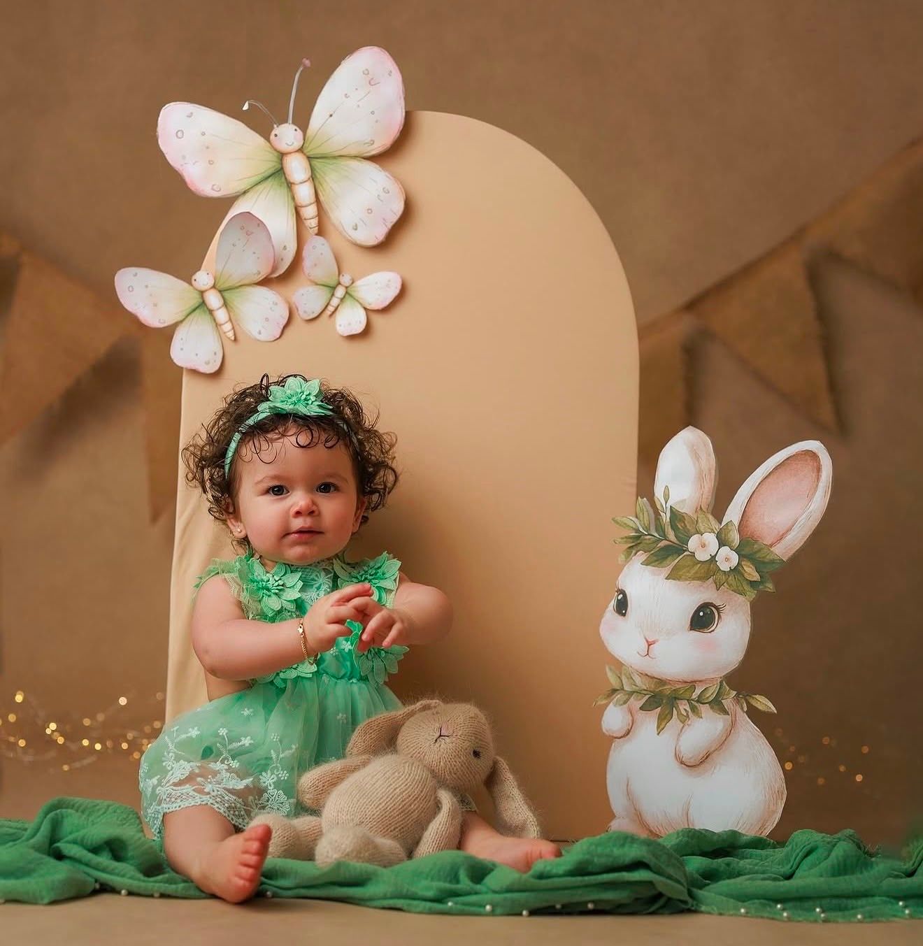 Baby girl in green lace outfit poses with decorative butterfly and bunny props in a charming Easter-themed photo session.