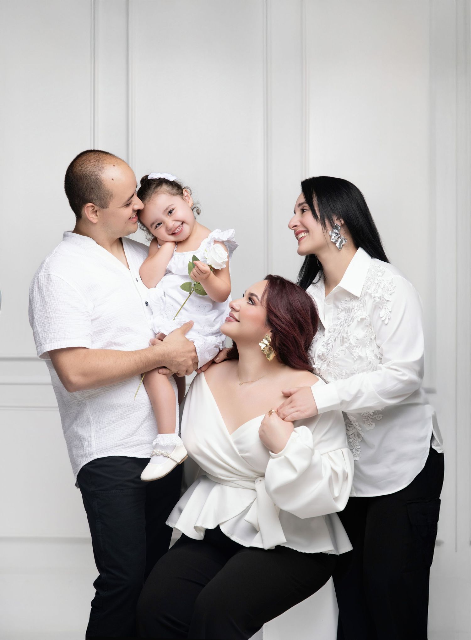 Family of four in white outfits posing joyfully against a white backdrop, parents smiling at toddler holding a flower.