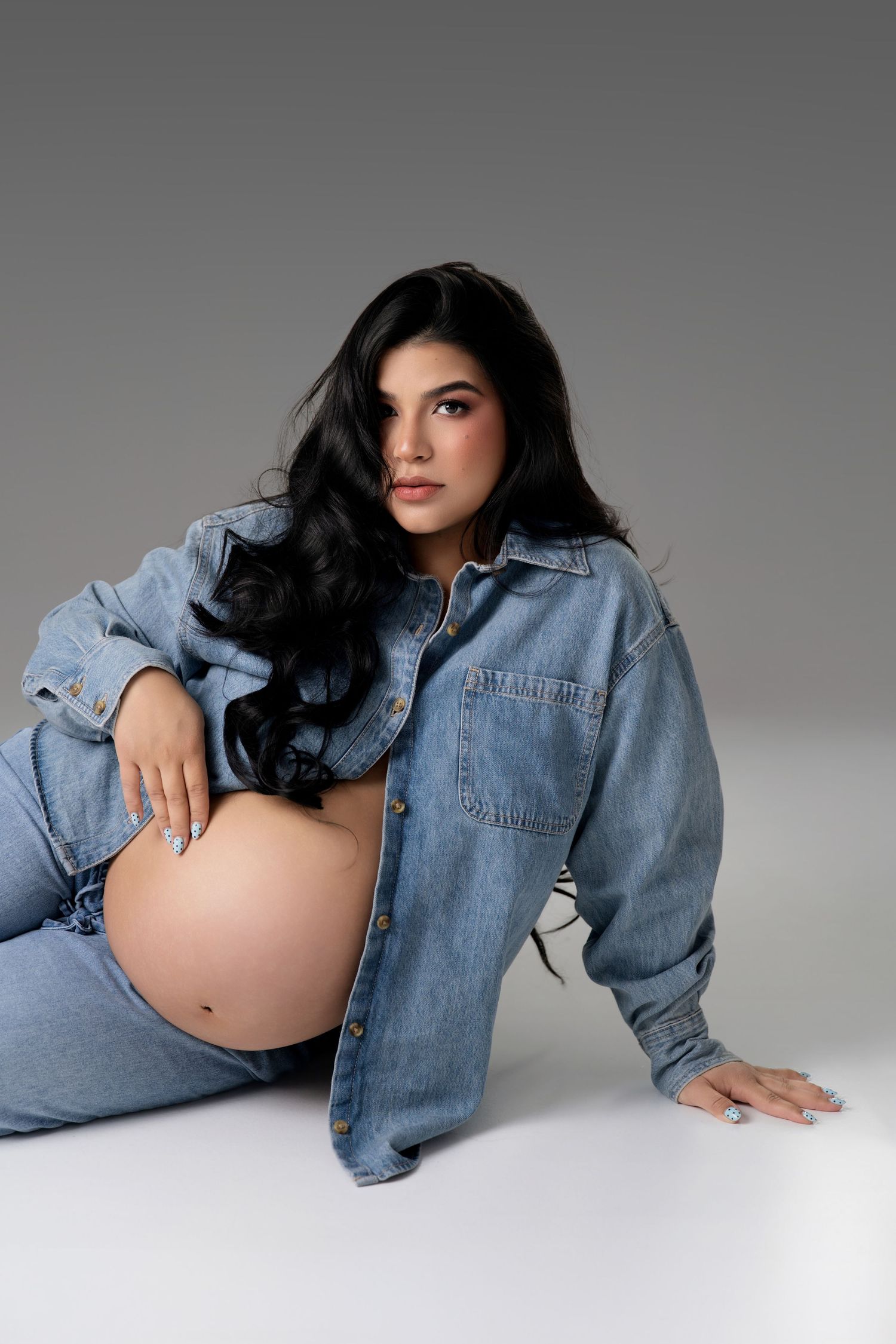 Pregnant woman in denim shirt and jeans sitting on gray background, cradling baby bump with long dark wavy hair.