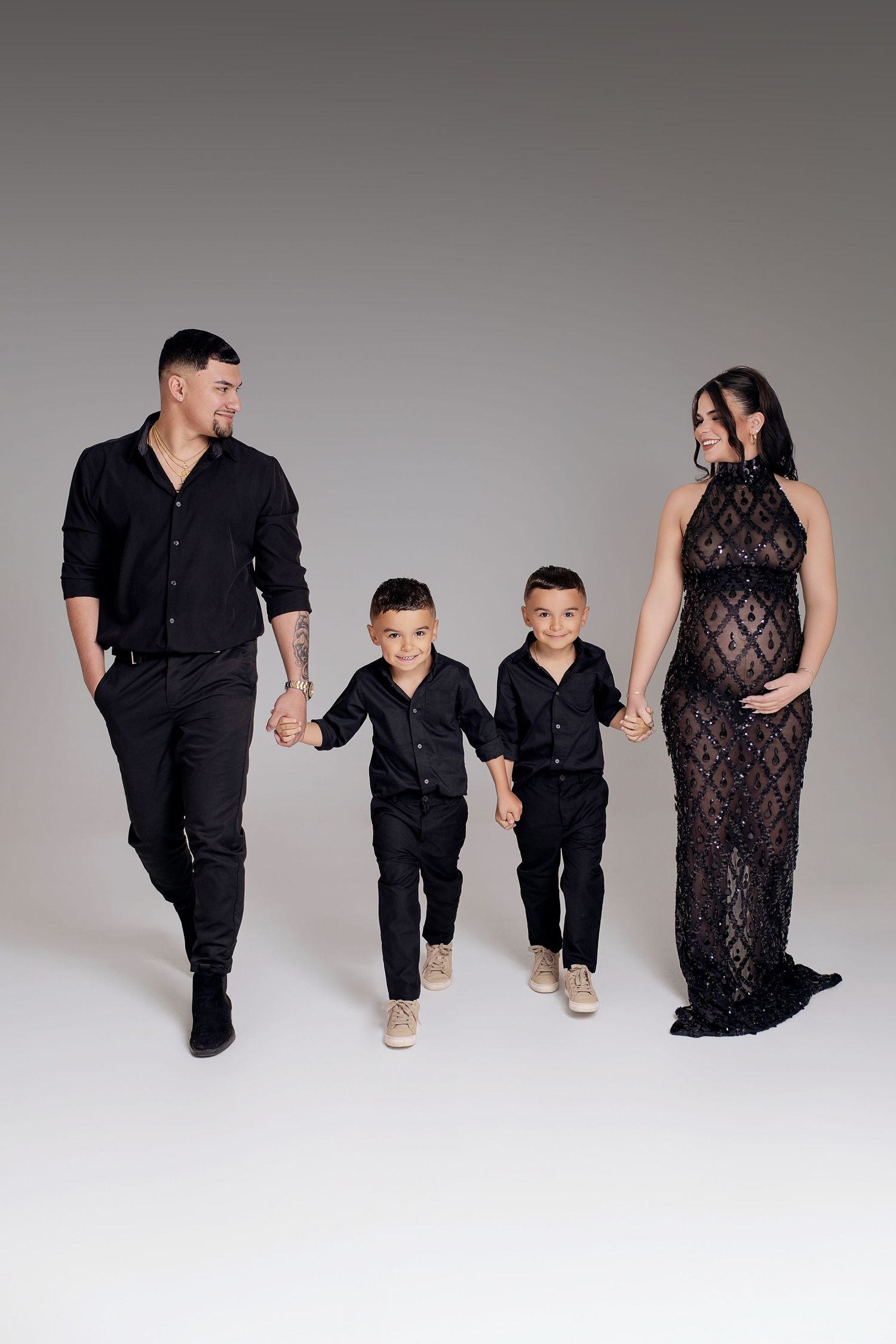 A family of four in matching black outfits, with a pregnant mother in lace gown, father, and two boys holding hands.