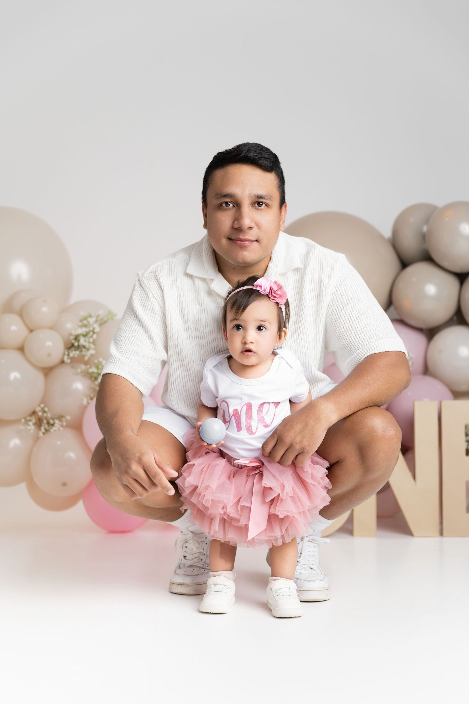 Father crouching behind baby girl in pink tutu skirt at first birthday party with balloon decorations.