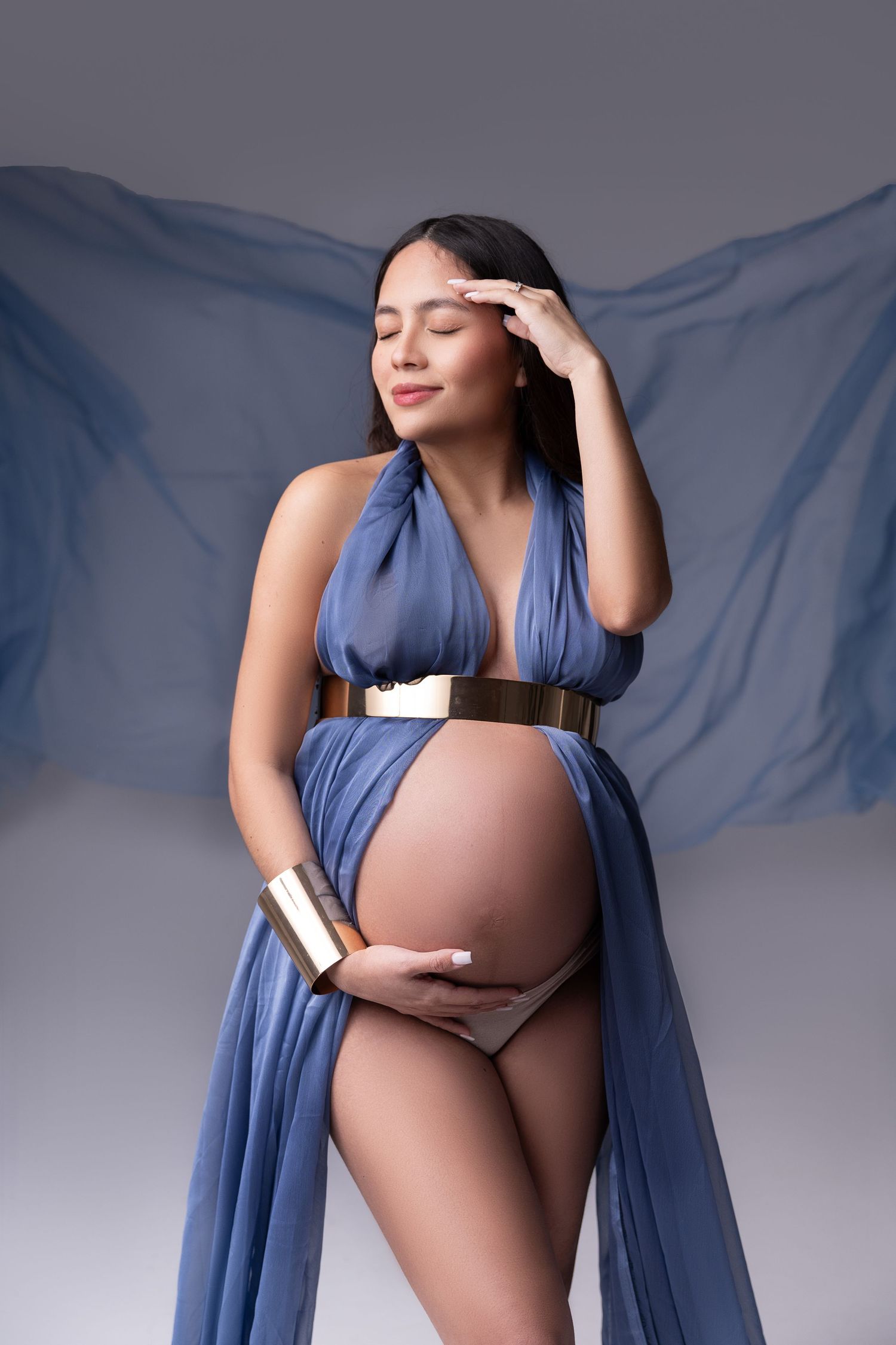 Pregnant woman in blue draped fabric dress with gold belt, cradling baby bump, posing gracefully against gray background.