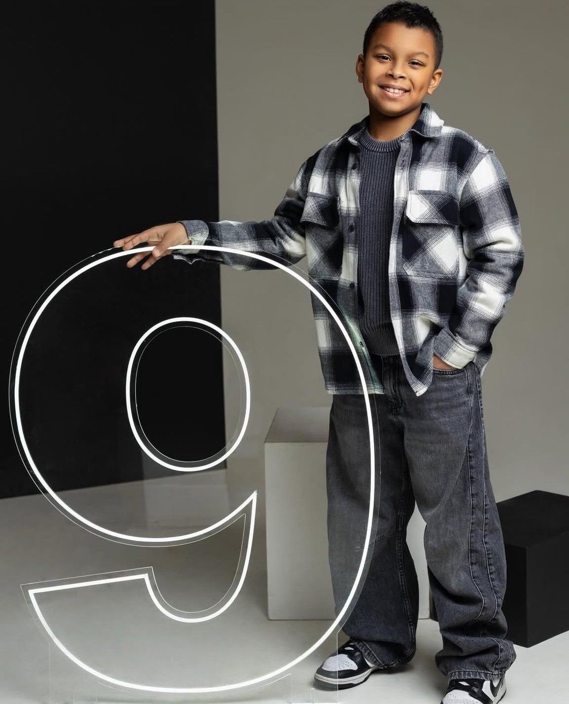 Young boy in plaid shirt posing with a large illuminated number 9 against a gray studio background.