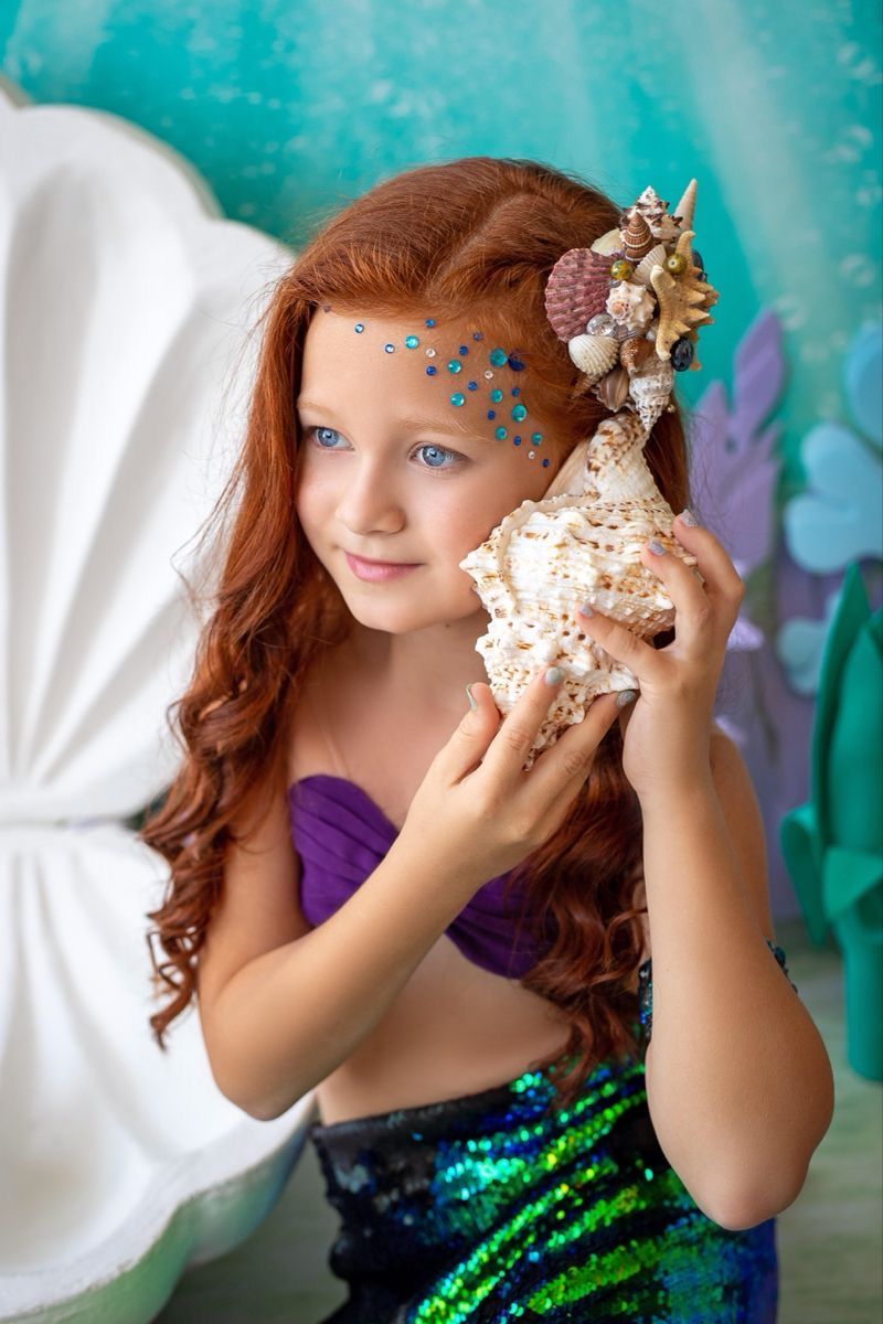 A young girl dressed as a mermaid with red hair, blue sequin skirt, shell accessories, holding a large conch shell.