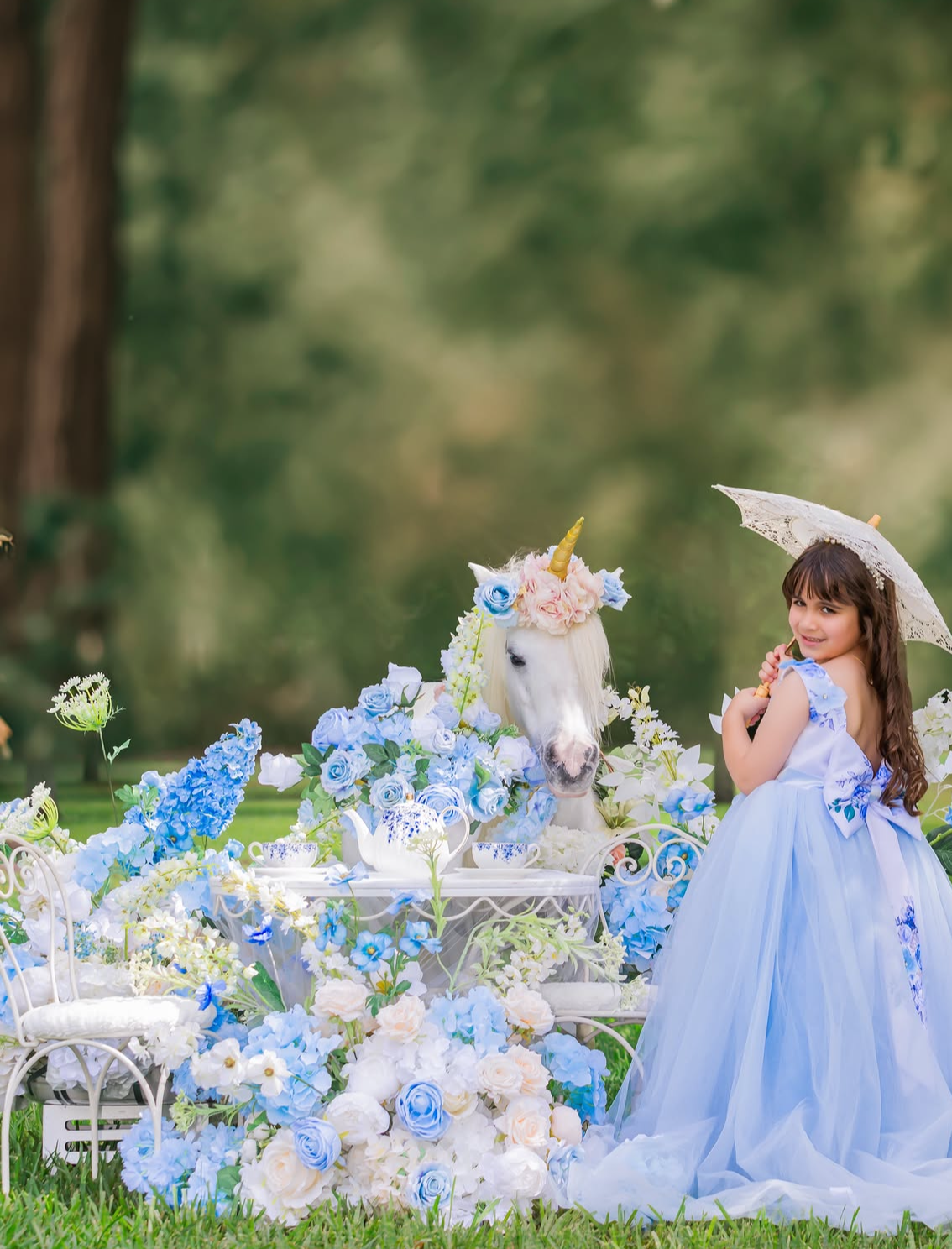 A young girl in a blue ball gown poses with a white unicorn amid blue and white floral arrangements outdoors.