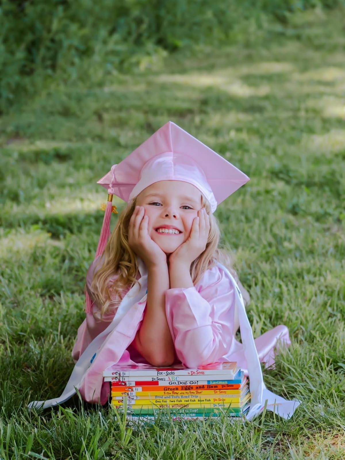 Young girl in pink graduation cap and gown lying on grass smiling with stacked colorful books.