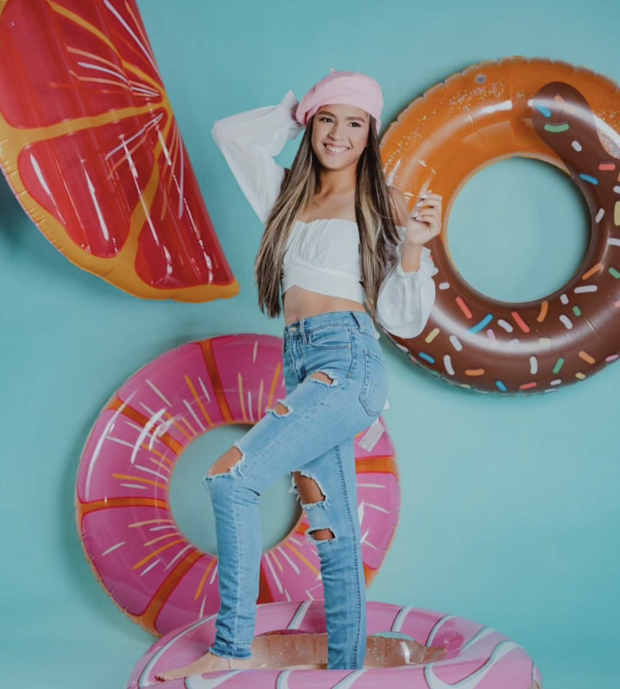 Young woman in white crop top and ripped jeans poses with colorful donut and fruit inflatable pool rings.