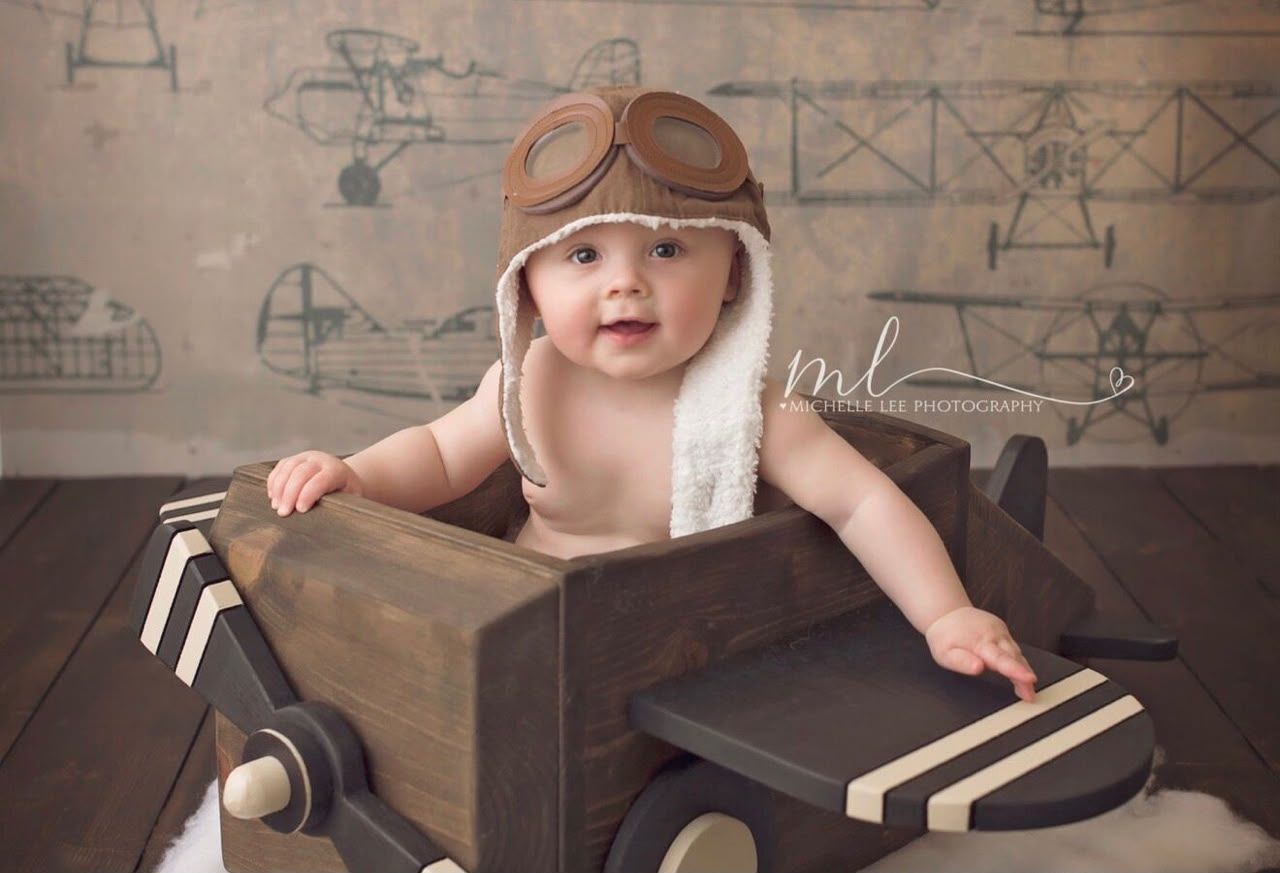 Baby in aviator hat and goggles sits in wooden toy airplane prop for a vintage-themed newborn photo session.