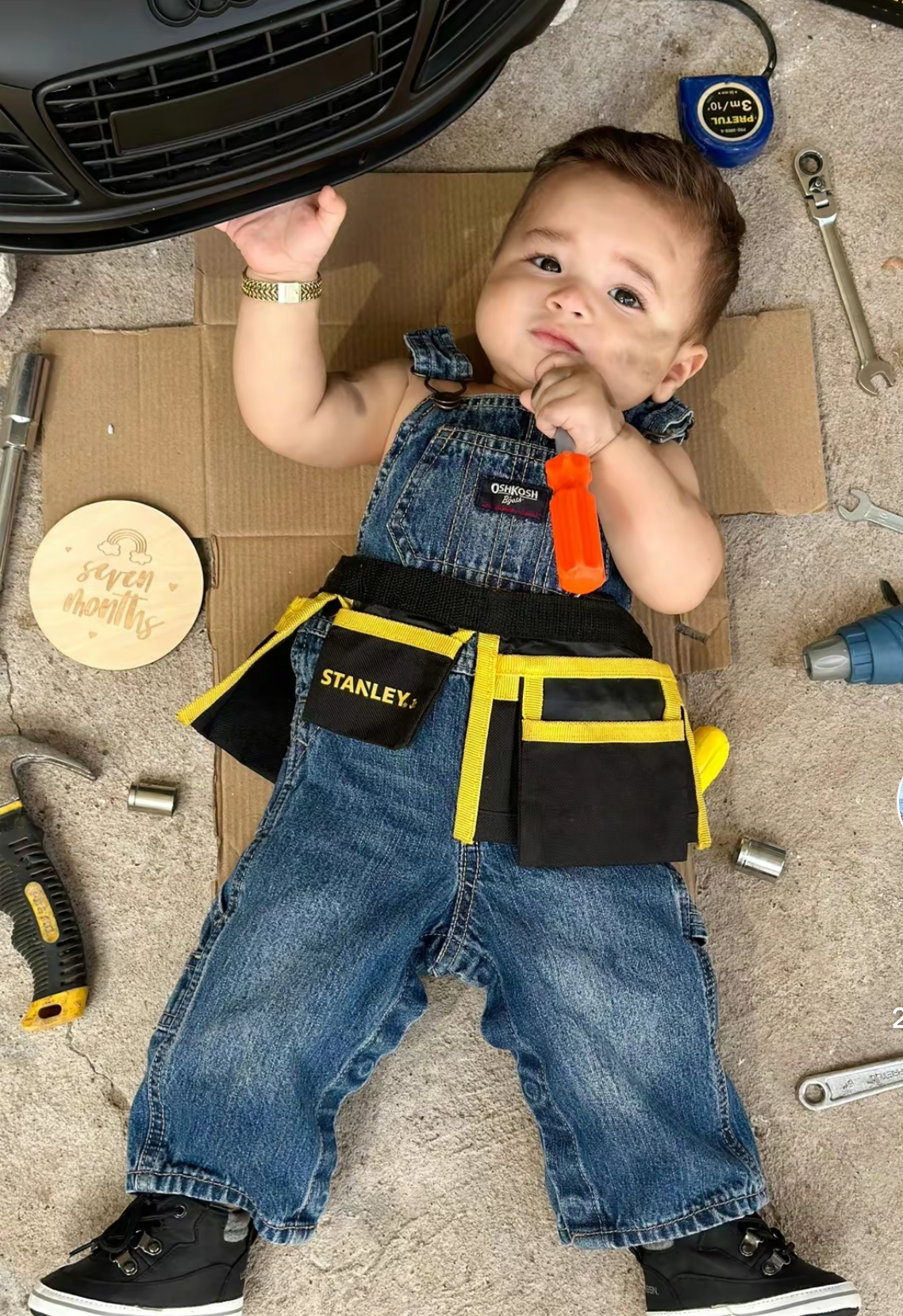 Adorable baby in denim overalls with Stanley tool belt, surrounded by tools, holding a tire in a cute seven-month photo shoot.