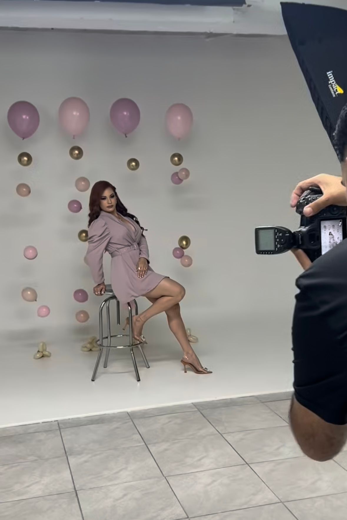 Woman in pink dress poses on stool during professional photo shoot with balloon backdrop and studio lighting.