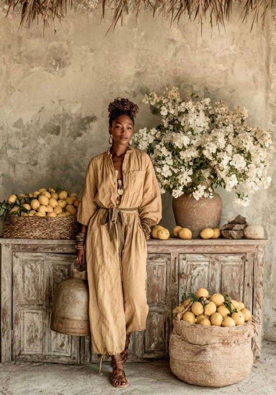 Woman in tan linen jumpsuit stands by rustic wooden dresser with lemons and white flowers in earthy Mediterranean setting.