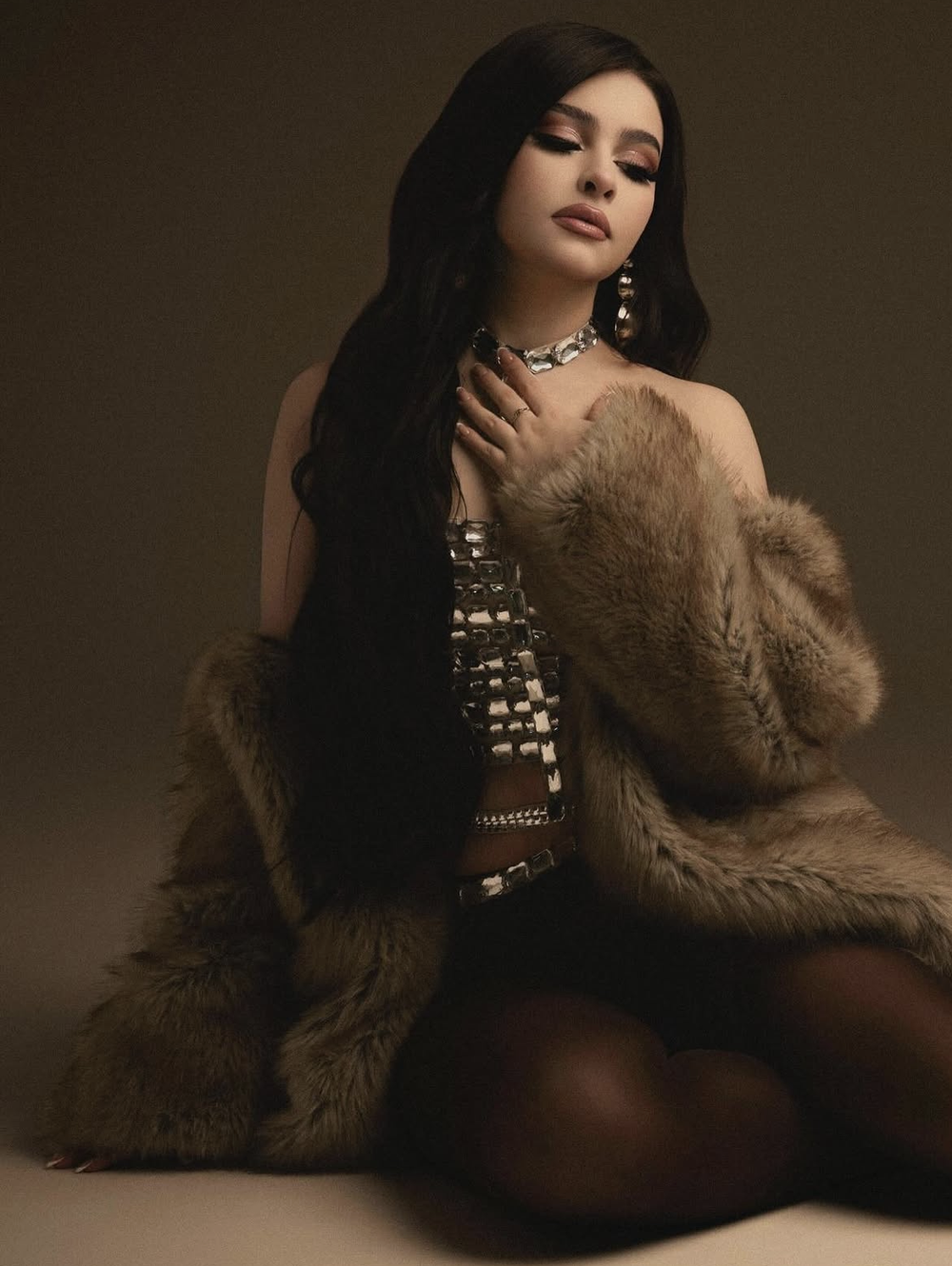 Young woman in luxurious fur coat and sequined dress poses gracefully with statement jewelry against dark background.