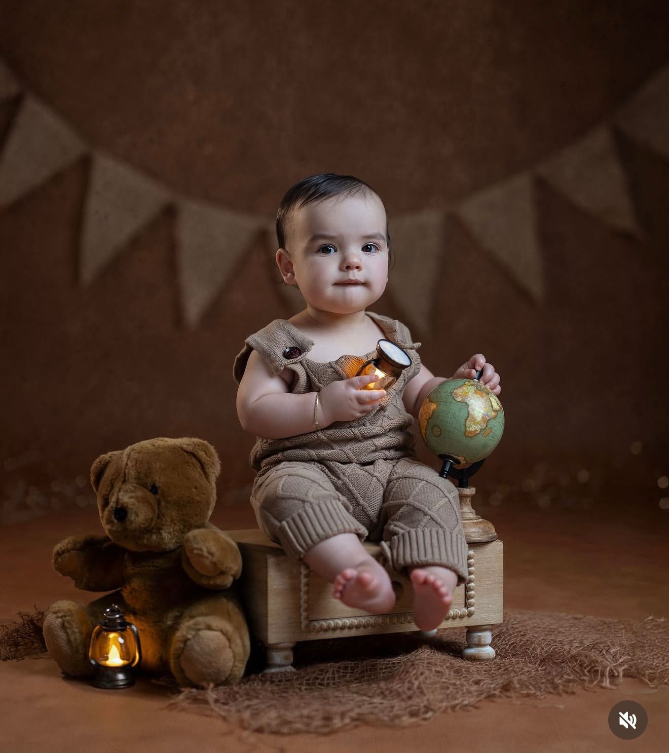 Adorable baby in knit outfit holds a globe, seated on a wooden crate with a teddy bear and lantern on a brown backdrop.