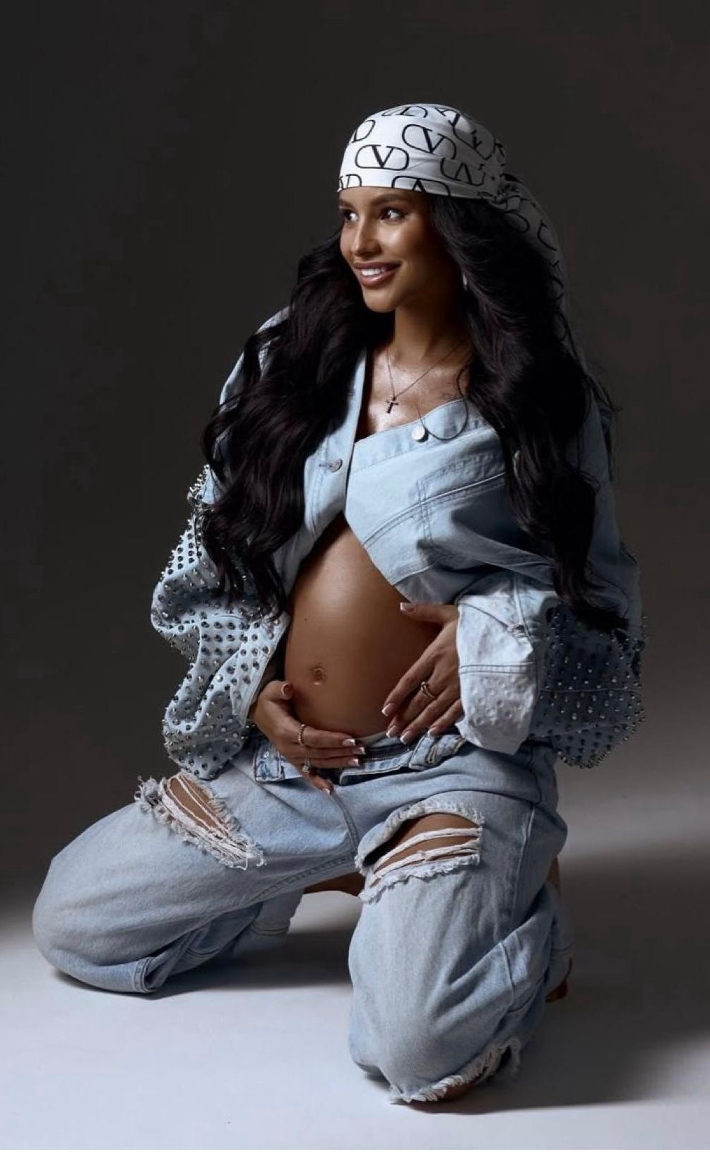 Pregnant woman in ripped denim jeans and designer bucket hat kneeling while cradling baby bump.