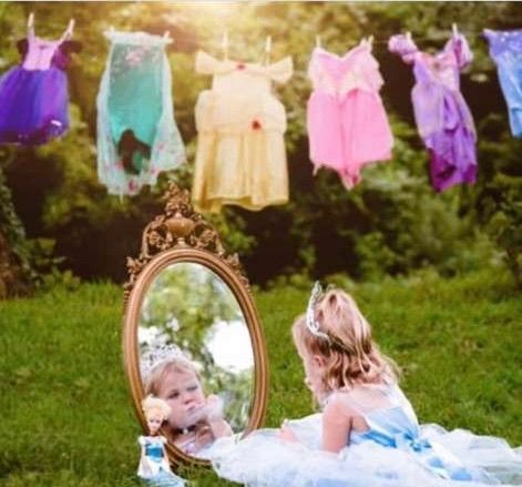 A little girl in a princess dress sits before a vintage mirror outdoors, with colorful princess dresses hanging on a line.