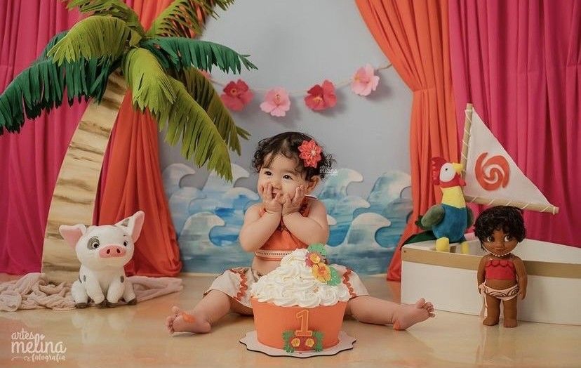 Baby girl sits before a Moana-themed cake smash backdrop with palm tree, ocean scenery, and character toys.